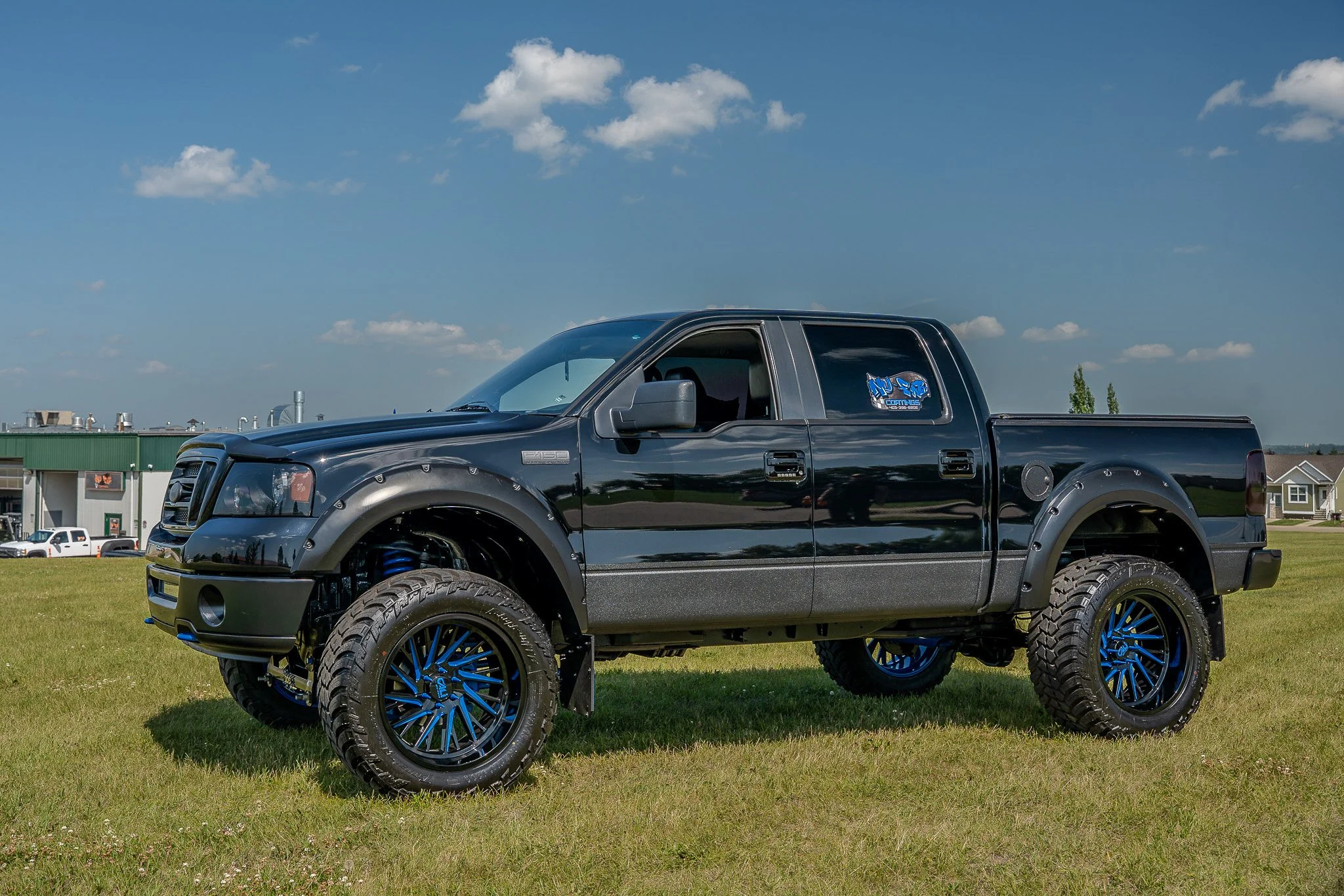Black lifted pickup truck with custom blue and black rims parked on grass under a partly cloudy sky.