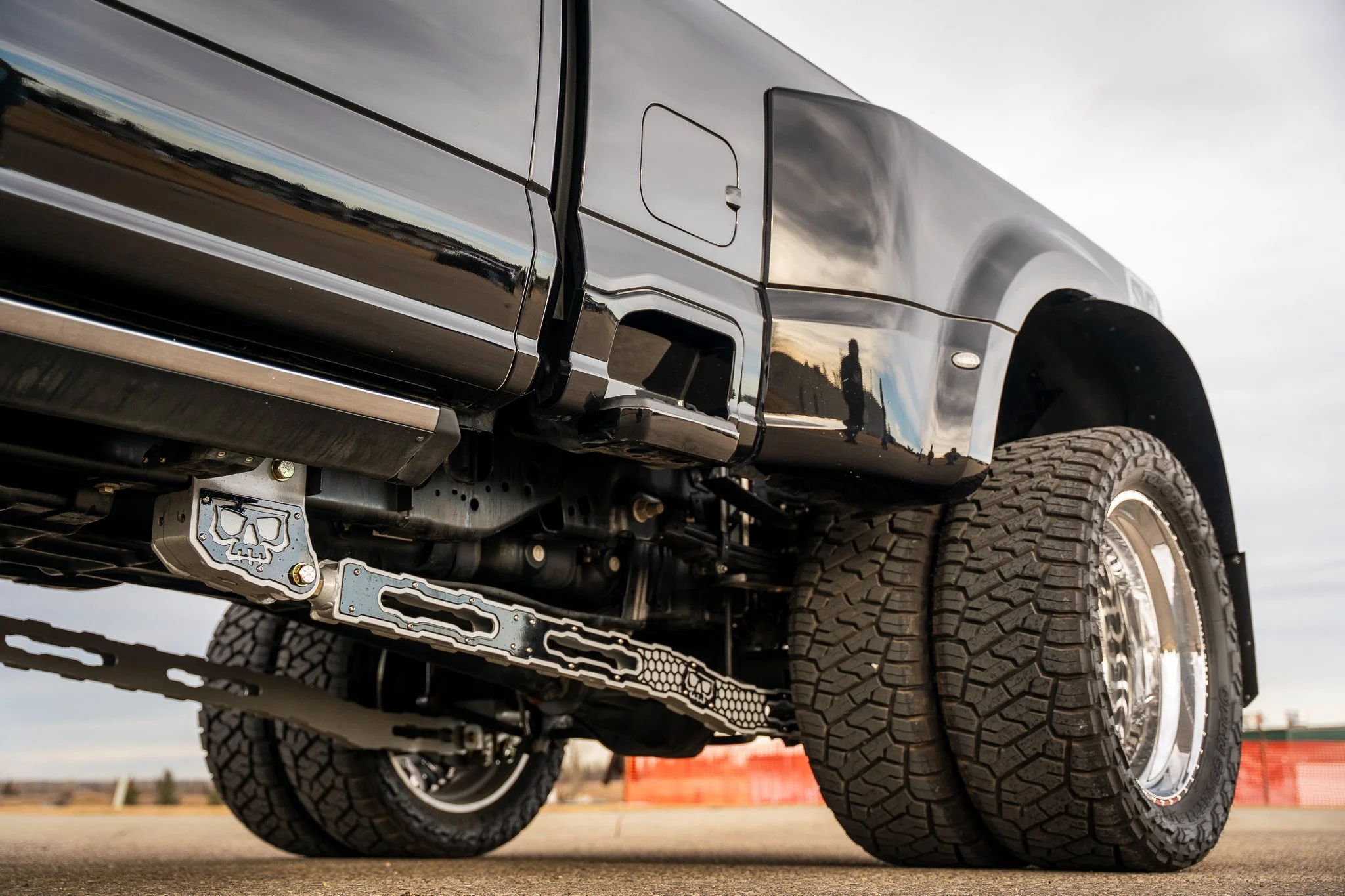 Close-up of lifted black truck with large off-road tires, custom suspension components with skull logo designs, parked on concrete with a cloudy sky.