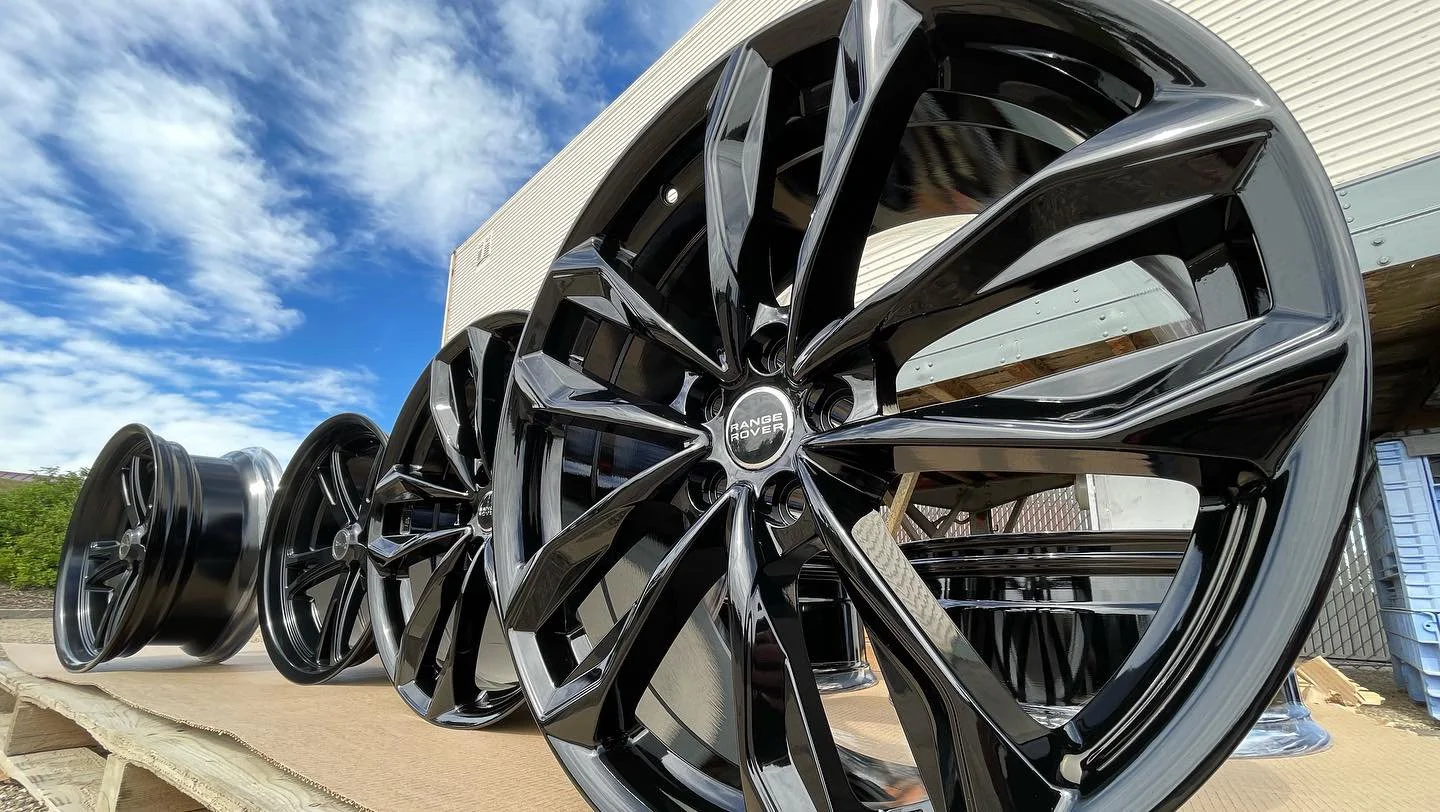 Multiple black alloy wheels displayed outdoors on a wooden surface with a building and blue sky in the background.