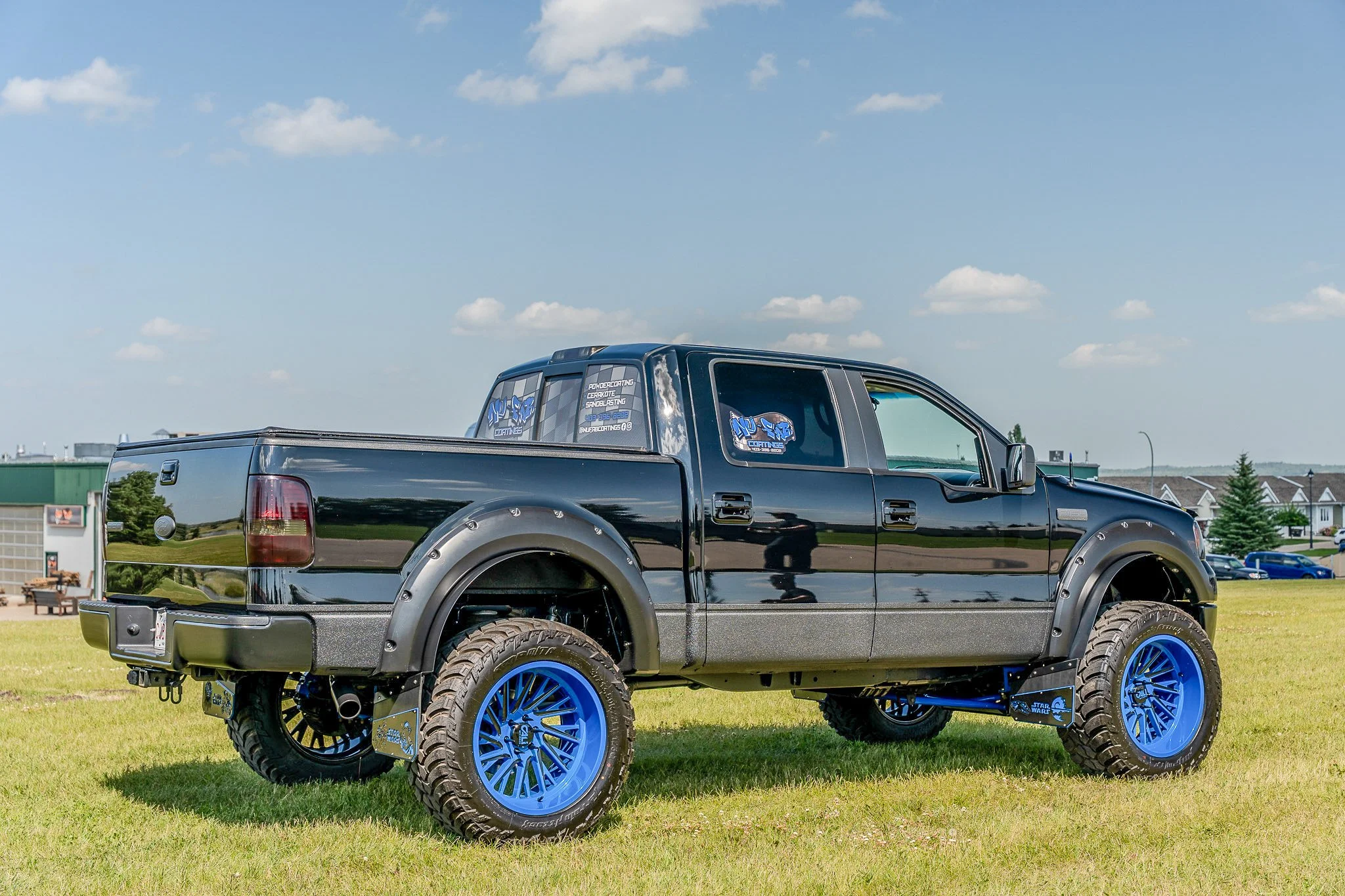Black pickup truck with blue wheels parked on grass, blue sky with clouds, residential buildings in the background.
