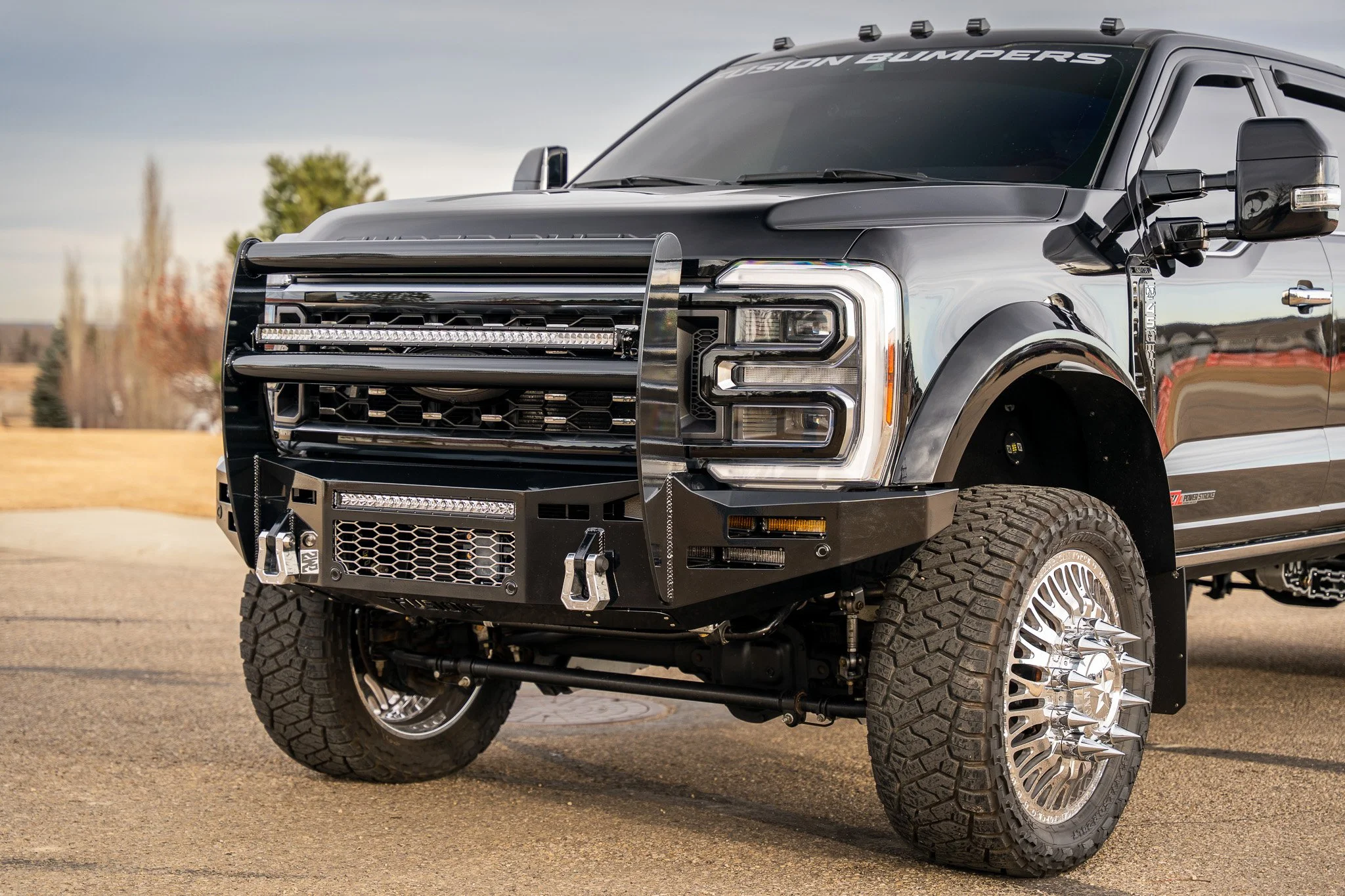 Black pickup truck with custom grille, off-road tires, and a heavy-duty front bumper, parked on pavement with a background of trees and sky.