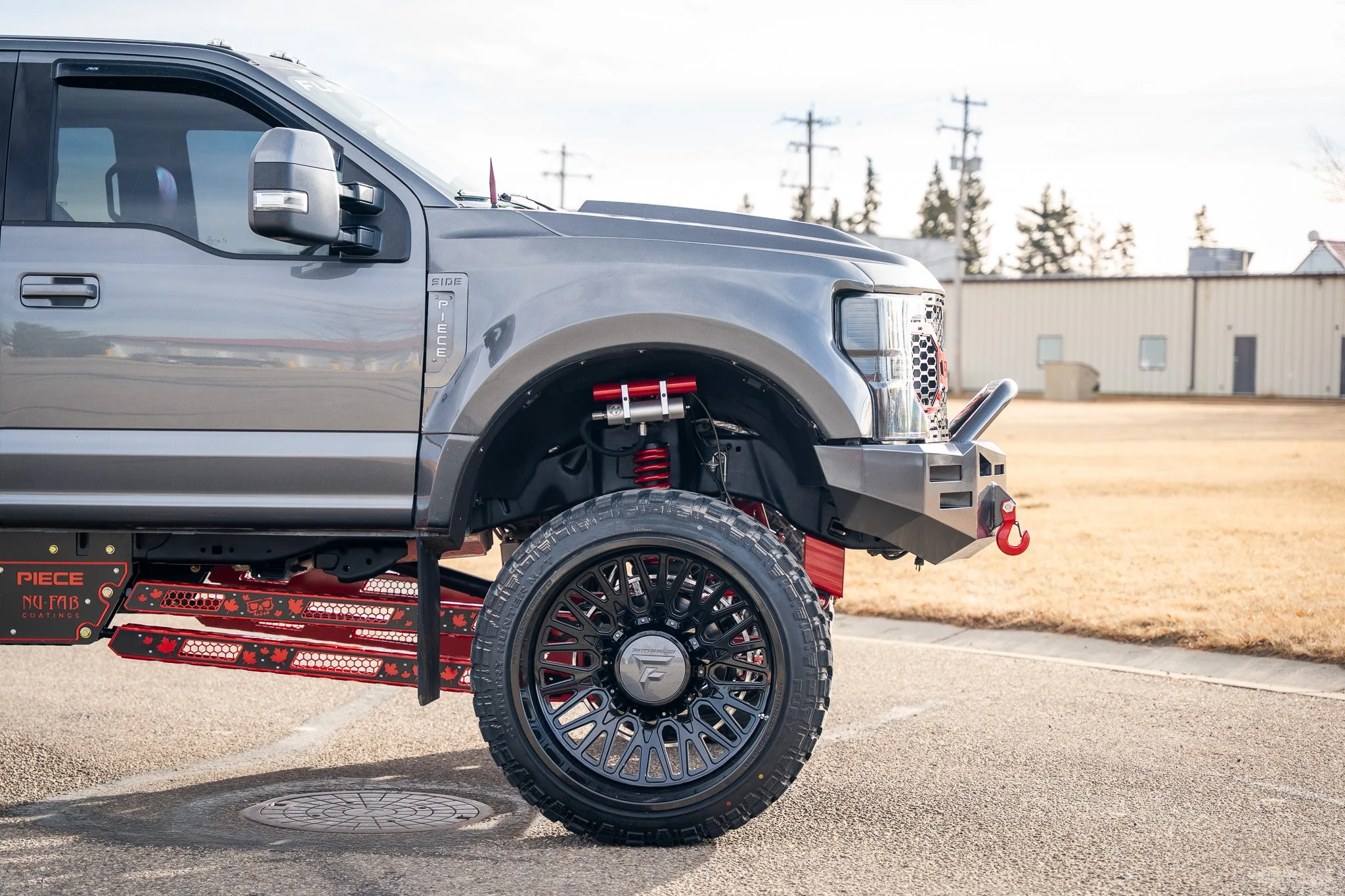 Close-up of a gray off-road pickup truck with red suspension parts and black aftermarket wheels, parked on a paved surface with a building and trees in the background.