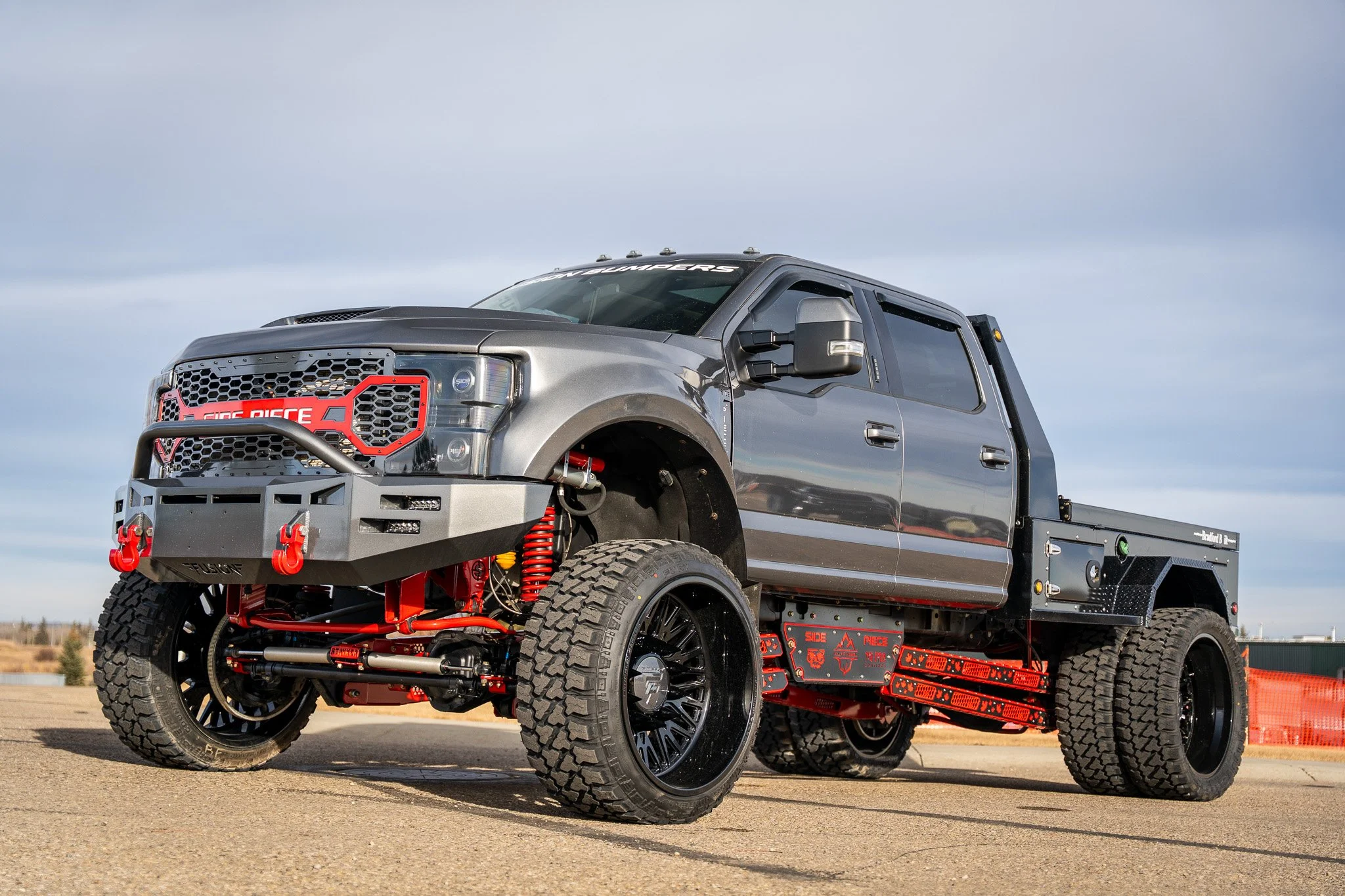 Custom modified truck with large off-road tires, gray body, black and red suspension components, and a flatbed in an outdoor setting.