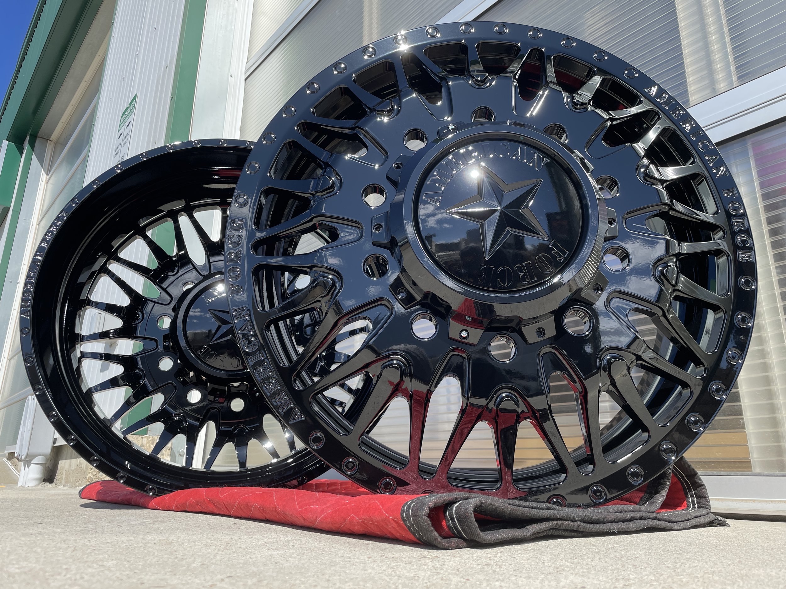 Two black, shiny, custom alloy wheels with star center caps, placed outdoors on a red mat in front of a building with sliding doors, reflecting clouds and sky in their surfaces.