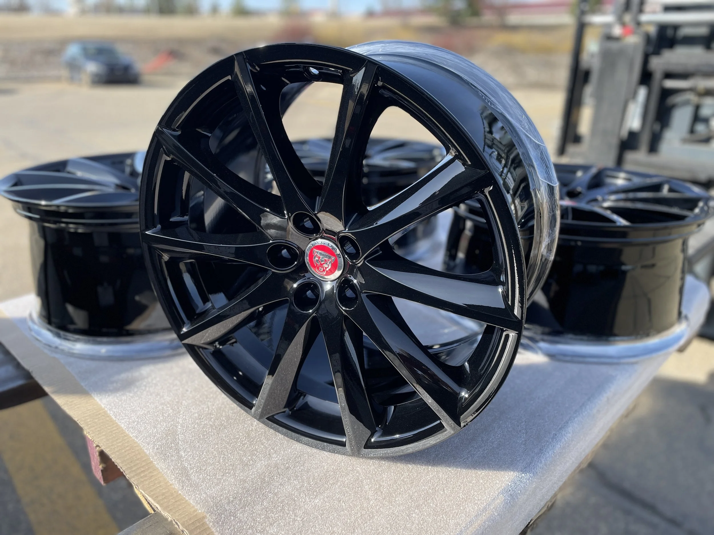 A stack of black alloy wheels, with one wheel in the foreground featuring a red and silver logo in the center, outside near a warehouse or industrial area.