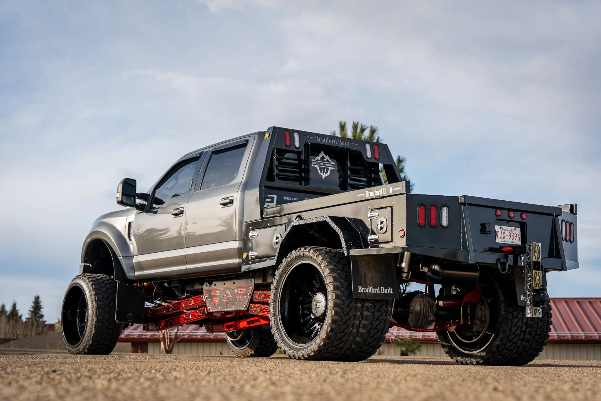 A custom-built pickup truck with a metallic silver cab and a flatbed, parked outdoors on a dirt surface, with a background of trees and a cloudy sky.