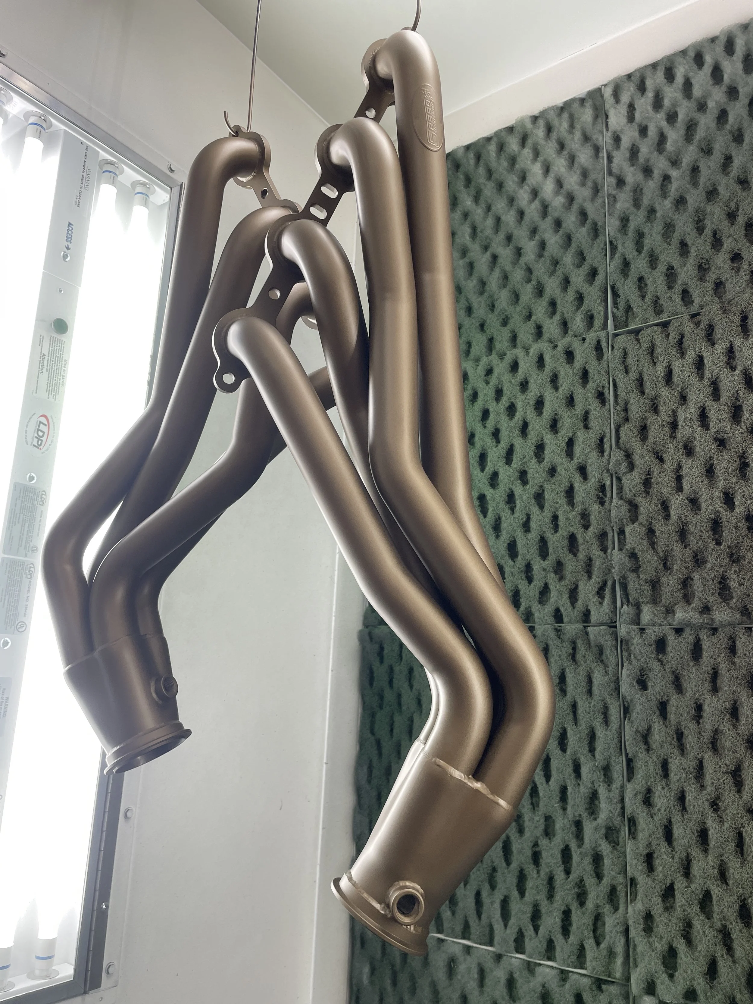 Four metallic pipes hanging from hooks in a room with black foam soundproofing panels on the wall.