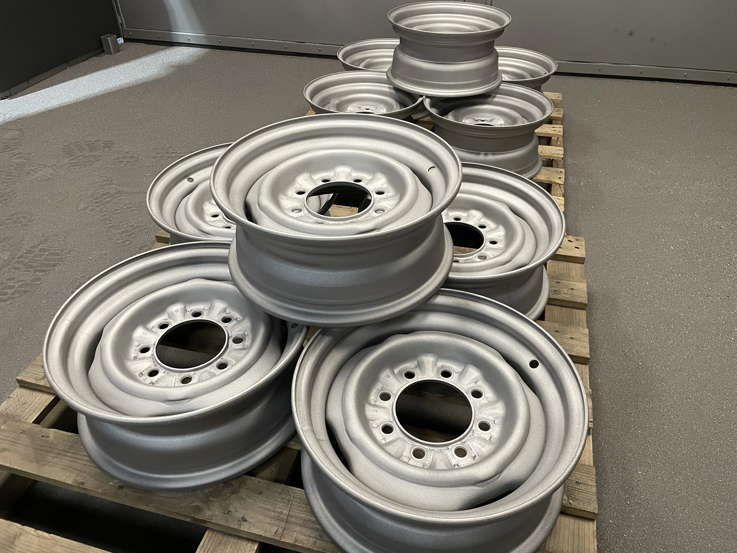 Multiple silver metal wheels stacked on a wooden pallet in a warehouse or workshop.