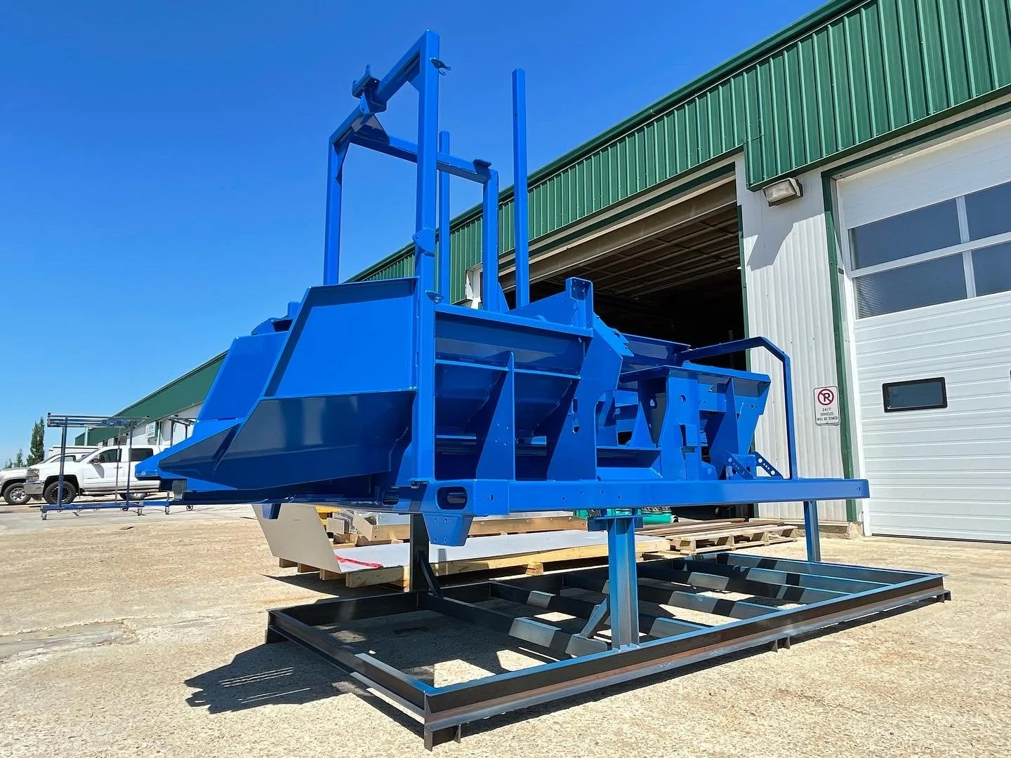 Large blue industrial or construction equipment frame outdoors near a green and white warehouse with a white door, parked vehicles in the background, under a clear blue sky.