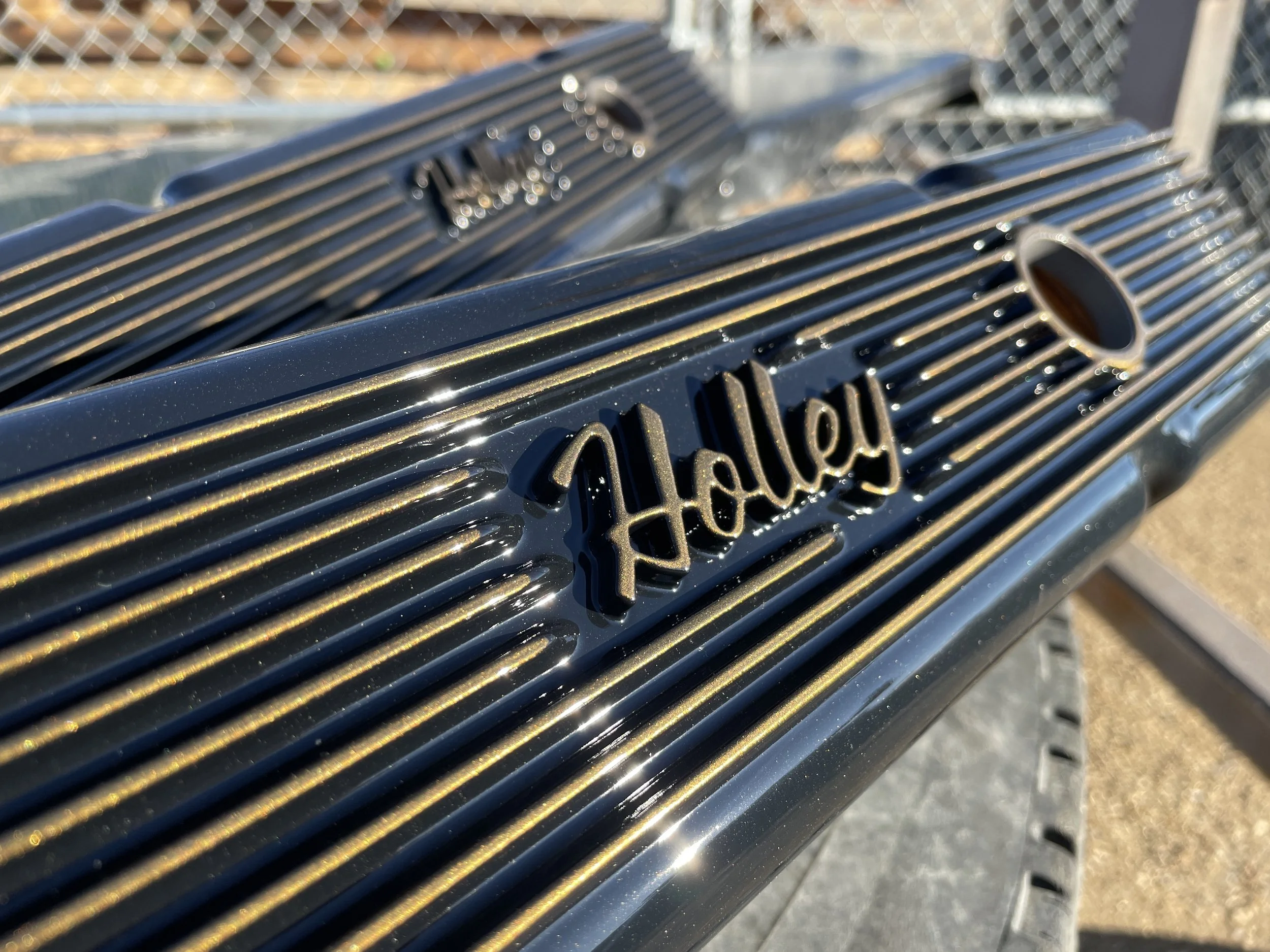 Close-up of a black Chevrolet valve cover with the name 'Holley' in raised letters, showing the cylinder fins and an oval opening for the ignition coil, with a blurred outdoor background.
