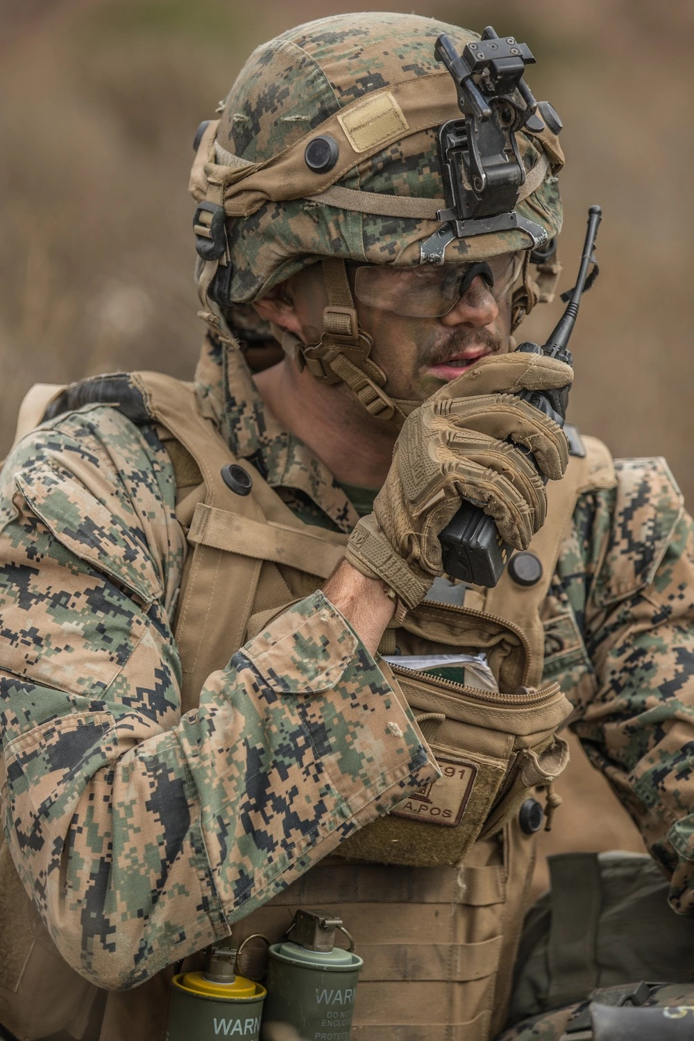 Marine in camouflage uniform talking on a radio, wearing a helmet, tactical gear, and sunglasses in an outdoor setting.