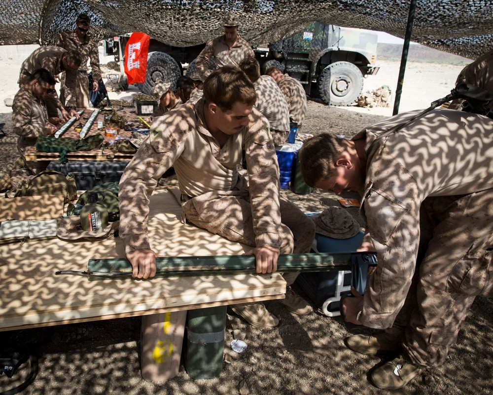 Marines in camouflage uniforms under a camo netting shade, working at tables with military equipment and supplies in a desert setting.