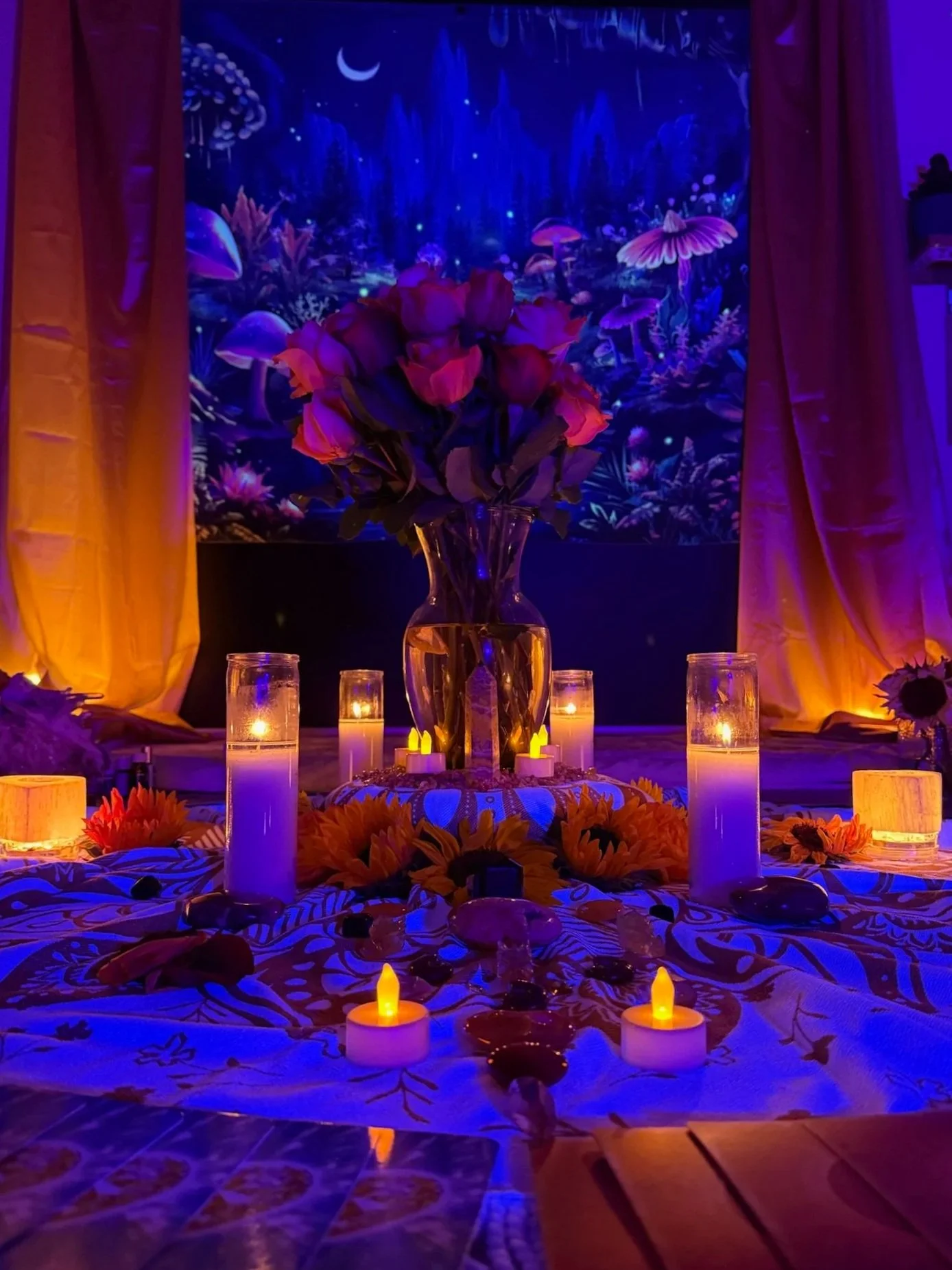 An indoor setting decorated with a large bouquet of pink roses in a vase, surrounded by candles, led light candles, and decorative stones on a table with a cloth. Behind the arrangement, there is a colorful, fantasy-themed backdrop with mushrooms, fl