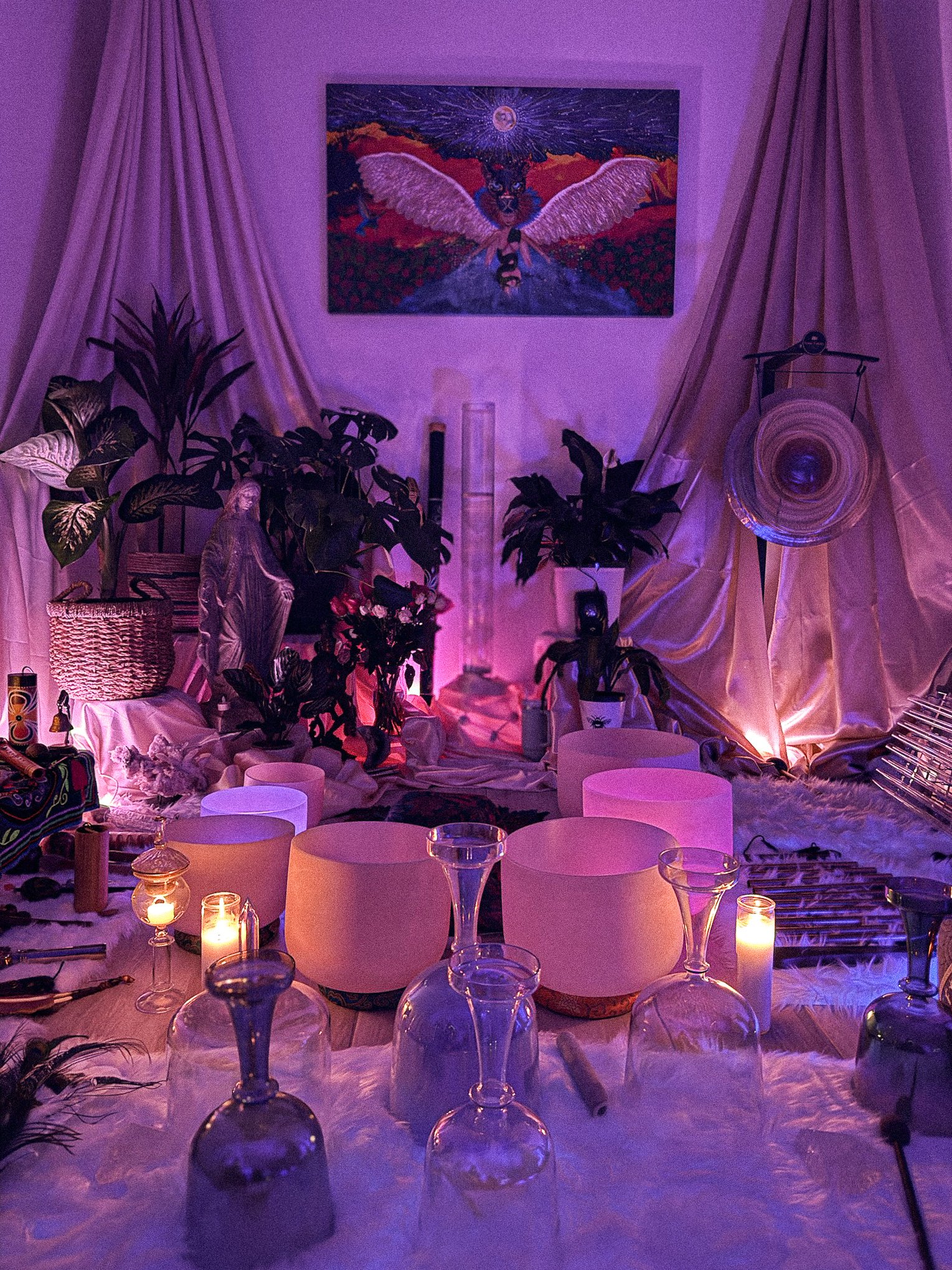 A cozy, dimly lit space decorated with plants, candles, and various musical instruments, featuring a painting of an angel with wings on the wall and draped curtains creating an intimate atmosphere.