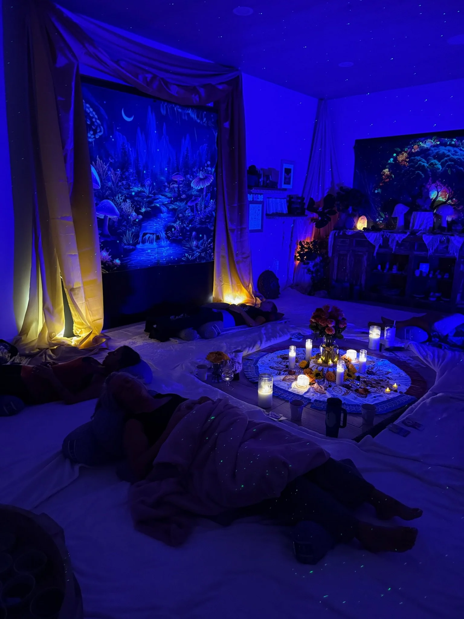 A cozy room decorated for a sleepover with starry-patterned ceiling, illuminated by candles and string lights, featuring a mural of a fantasy landscape with mushrooms and waterfalls, and a central arrangement of flowers and candles on a mat.