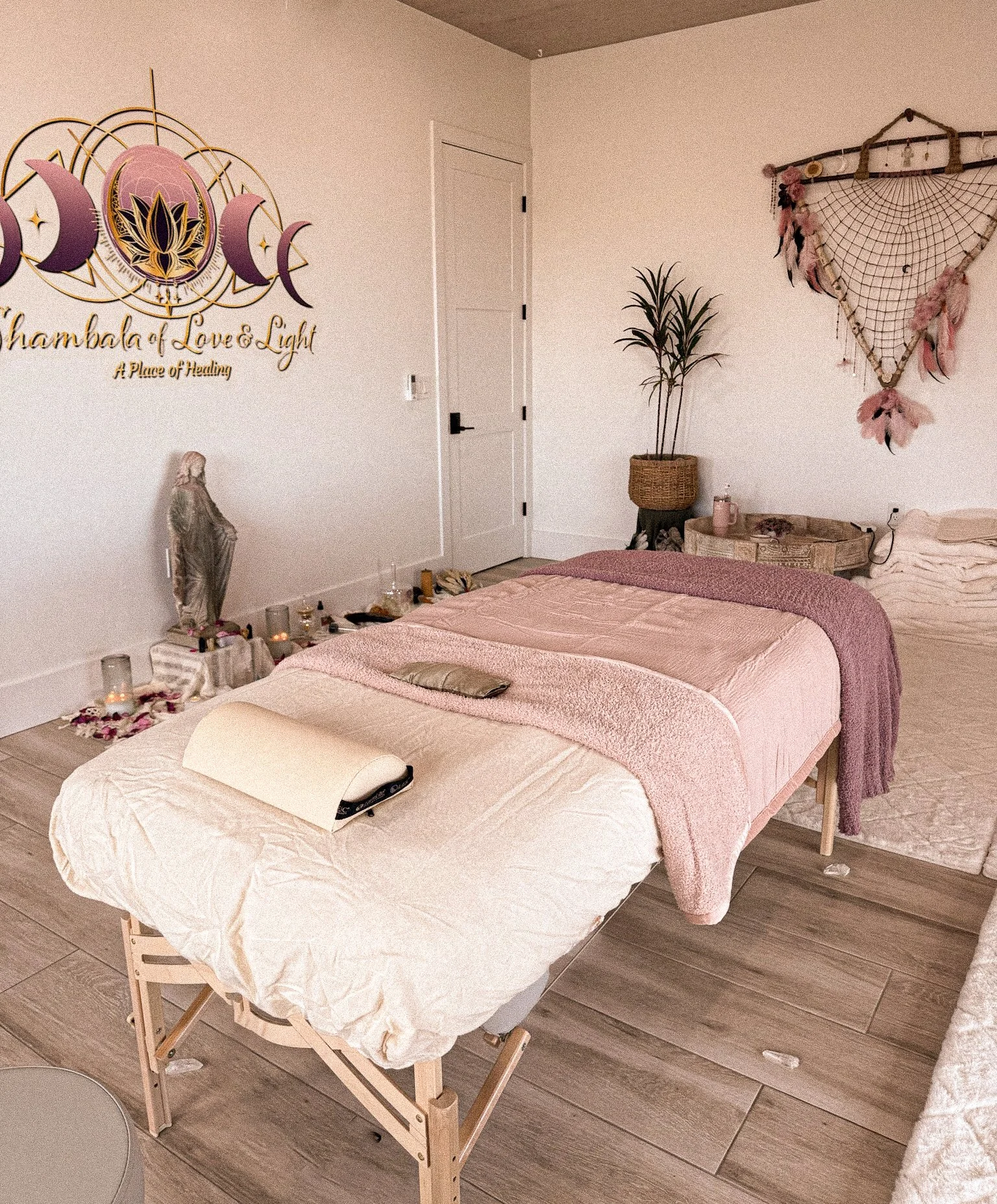A massage therapy room with a massage table covered in beige and pink blankets, a small statue, candles, and rose petals near the wall. The wall has a decorative sign reading "Shambala of Love & Light," featuring moon phases and a lotus flower. There is a doorway, a potted plant, and wall hanging with feathers and netting in the room.