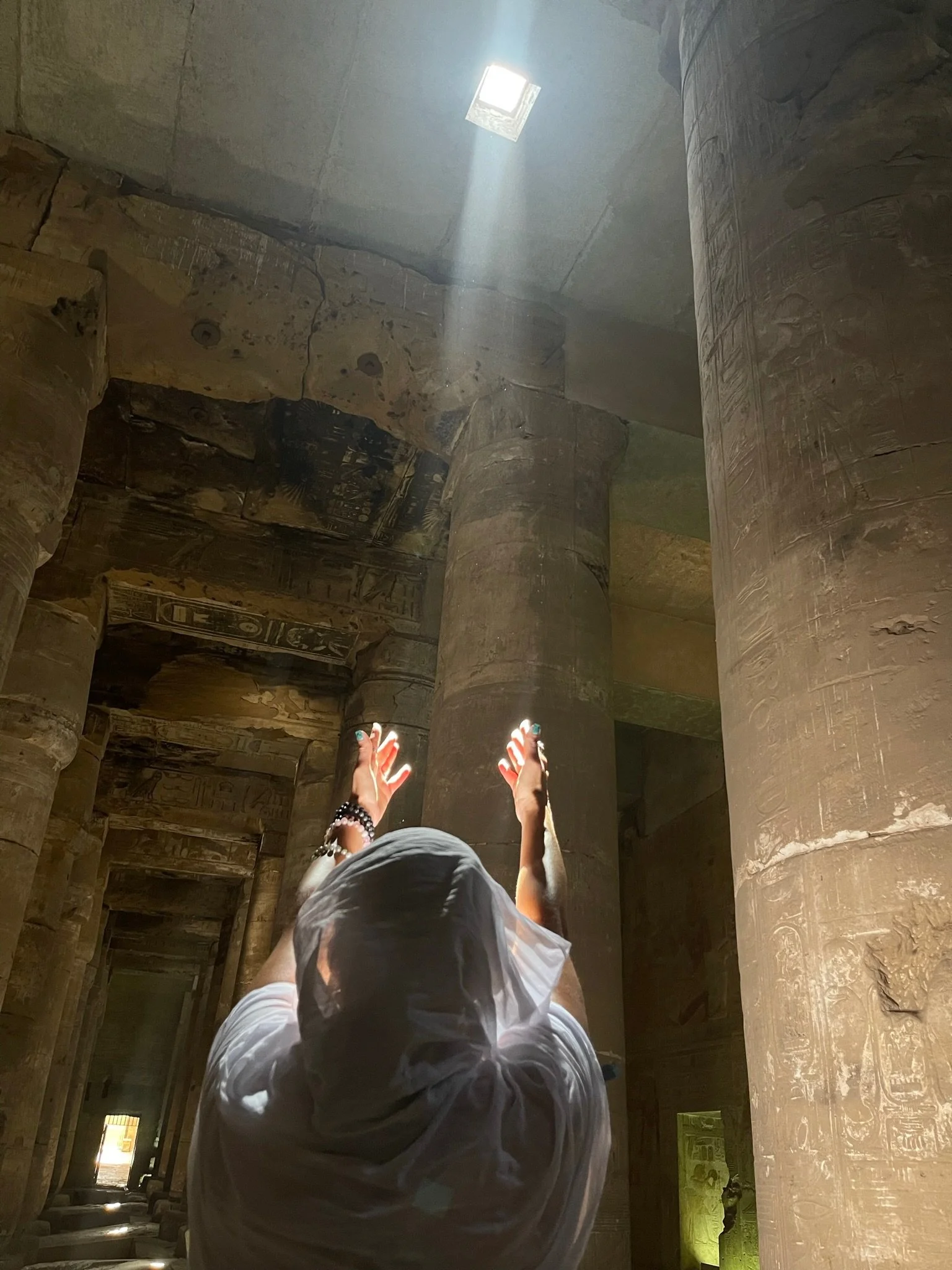 Person with head covered and arms raised inside an ancient Egyptian temple, illuminated by a beam of sunlight through a skylight, surrounded by large hieroglyphic carved stone columns.
