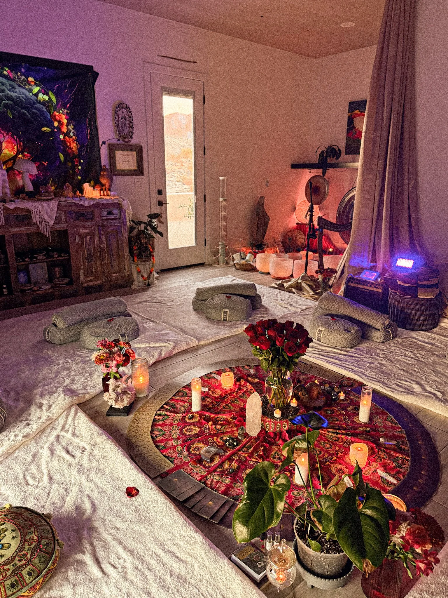 A cozy room decorated with candles, flowers, and spiritual objects for a ritual or meditation session. The room has soft lighting, cushioned seating, and various decorative items on shelves and tables.
