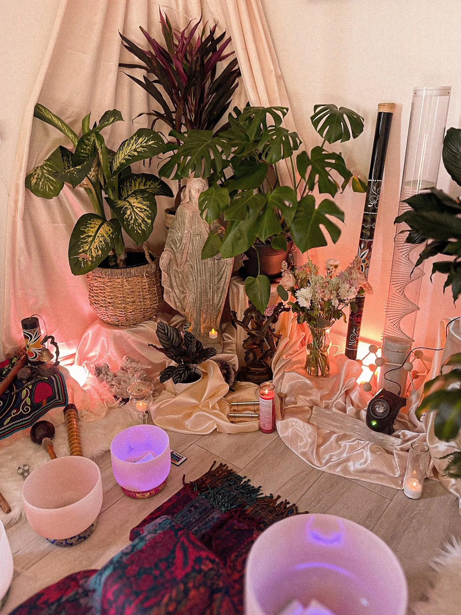 Indoor altar setup with large potted plants, candles, crystals, flowers in a vase, a statue, and decorative items on a draped fabric background with soft lighting.