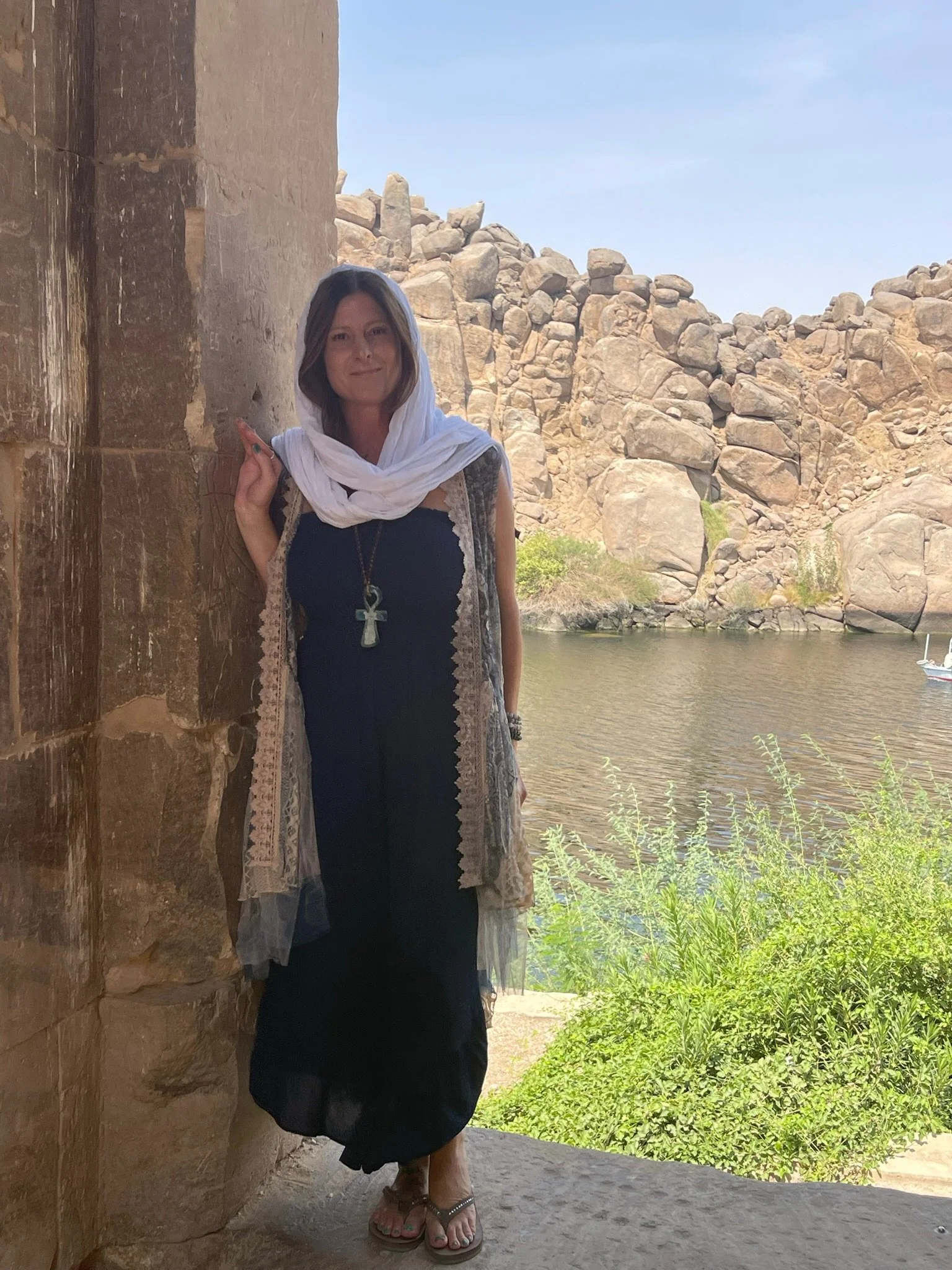 A woman dressed in black with a white headscarf, standing beside an ancient stone structure near a river with rocky hills in the background.