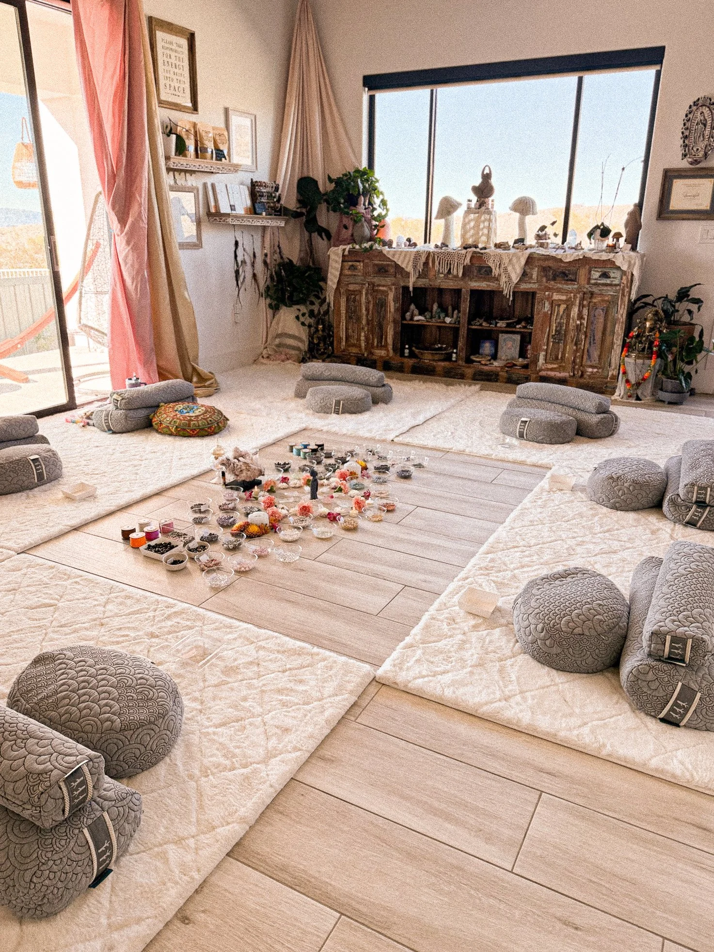 A cozy meditation room decorated with cushions and rugs, featuring a central wooden floor area with small bowls and offerings, large windows, and a rustic sideboard with various decorative items and plants.