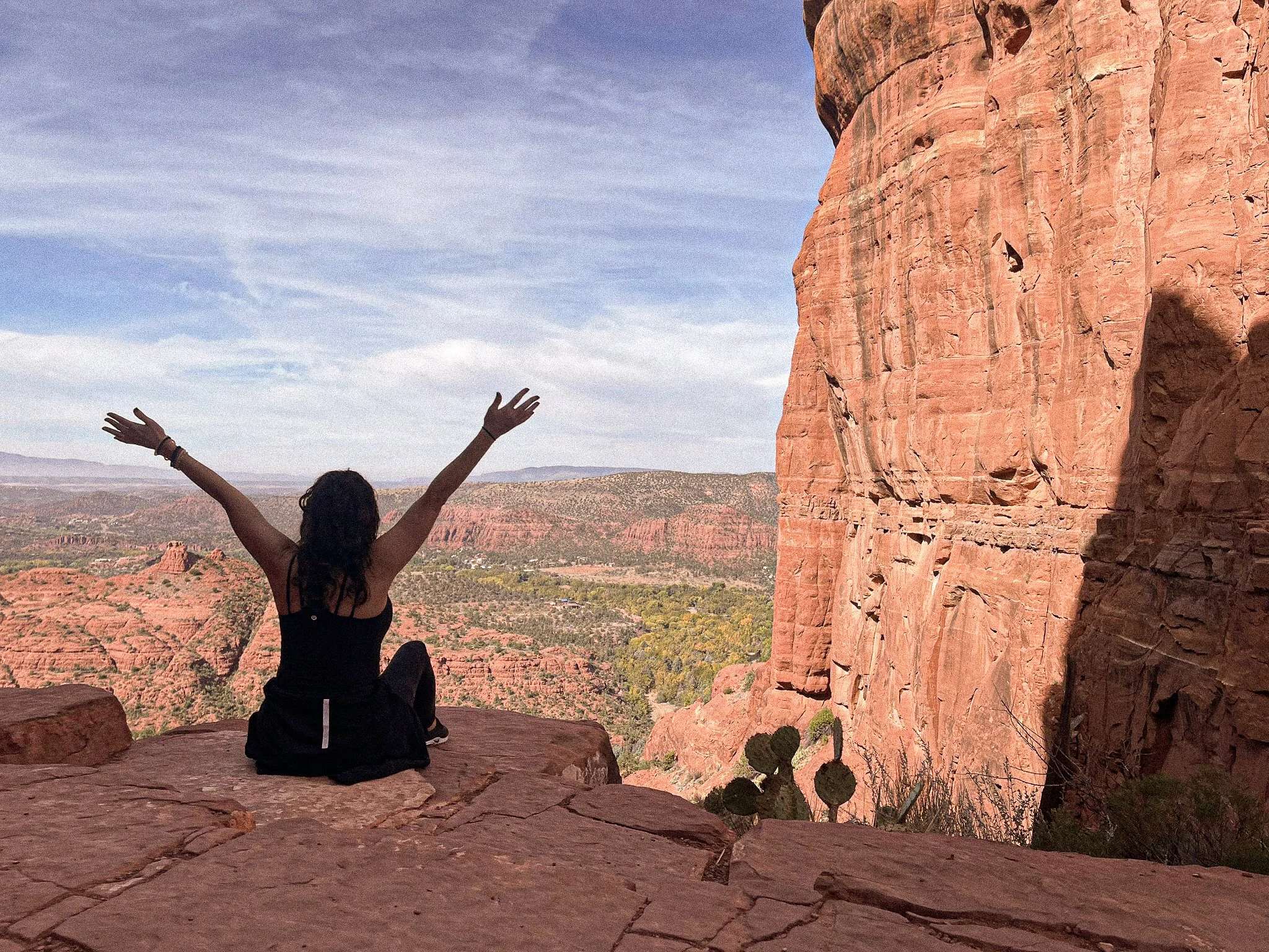 Person with long dark hair sitting on a large rock with arms raised, overlooking a desert canyon with red rocks and sparse vegetation under a blue sky.