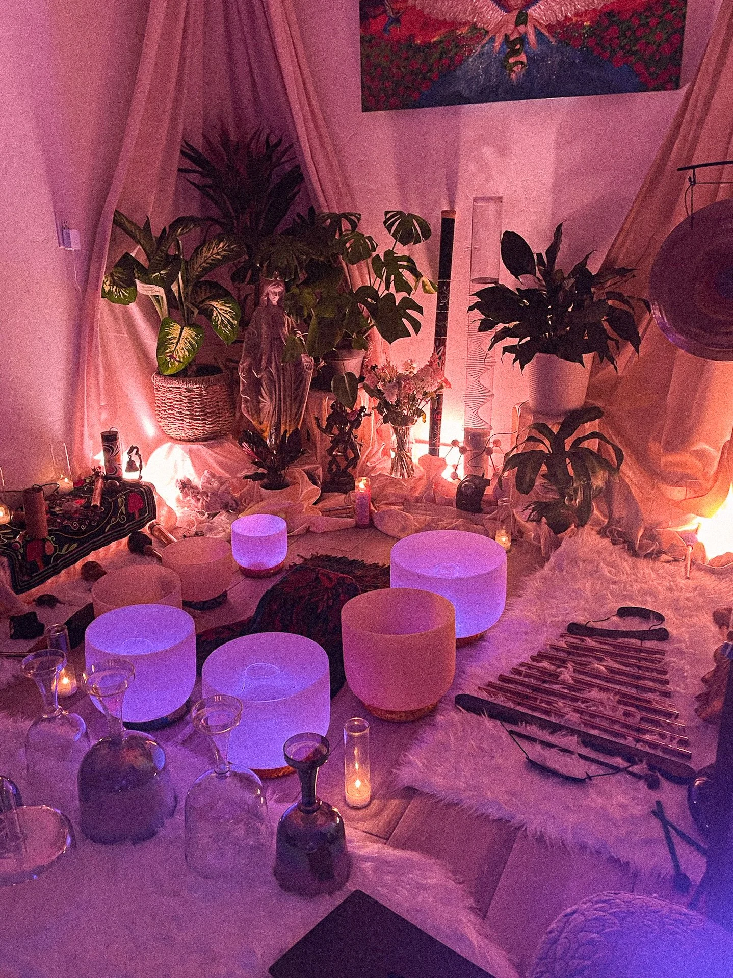 A cozy, dimly lit space decorated with plants, candles, and chimes, featuring pink and purple lighting, a painting of a bird on the wall, and a white furry rug.