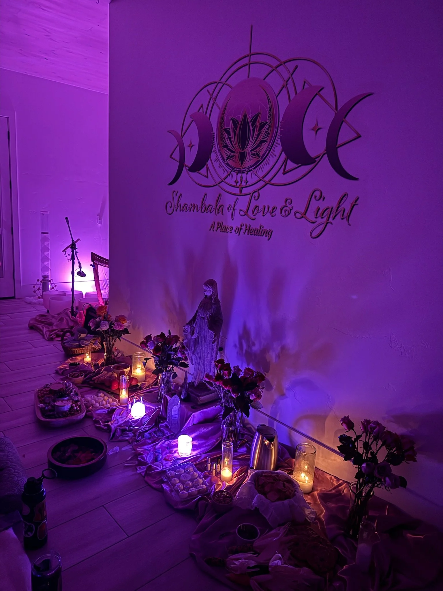A spiritual altar with candles, flowers, food, and religious figurines, illuminated with purple lighting. A large wall sign reads 'Shambala of Love & Light, A Place of Healing.'