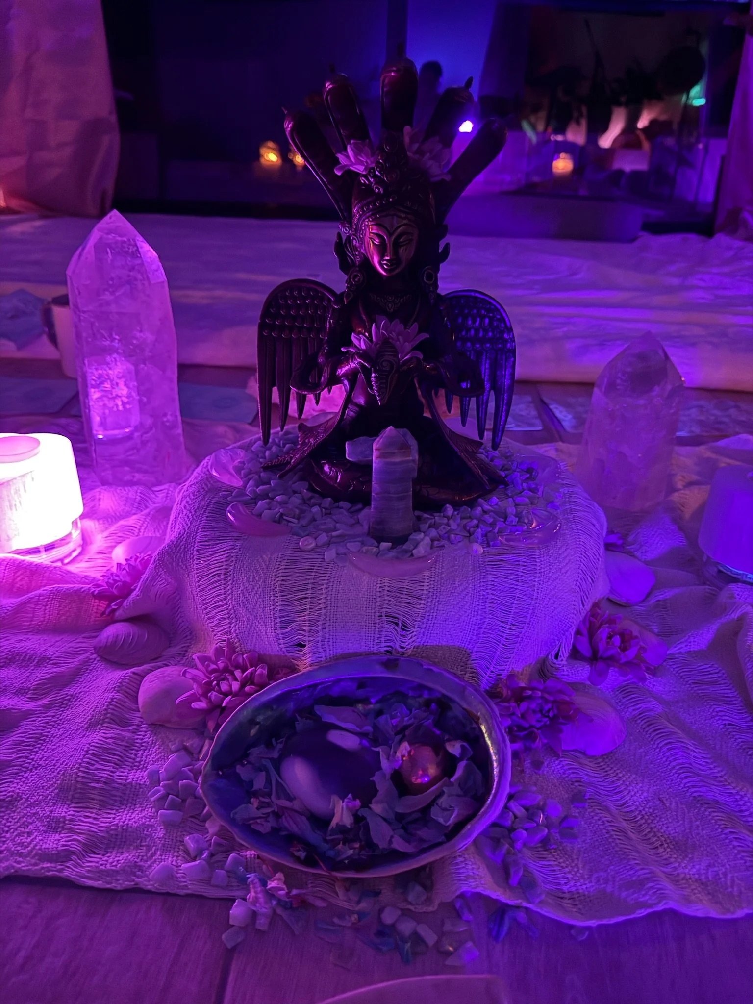 A spiritual altar with purple lighting, featuring a central statue of a deity with wings and a headdress, surrounded by crystals, shells, candles, and flowers on a cloth.