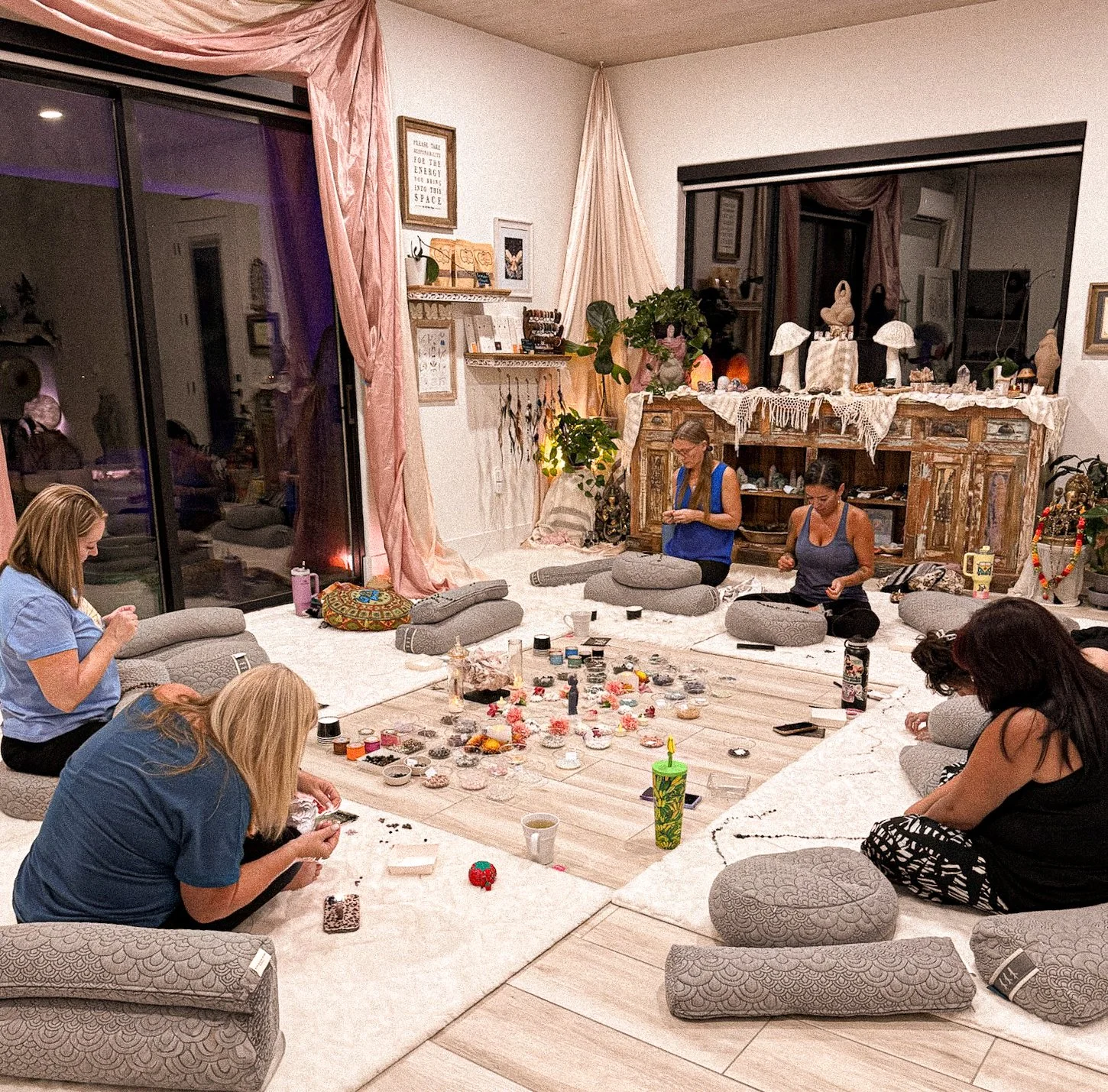 A group of women sitting on cushions in a circle on the floor, participating in a crafts or jewelry-making activity in a cozy, well-decorated living room at night.