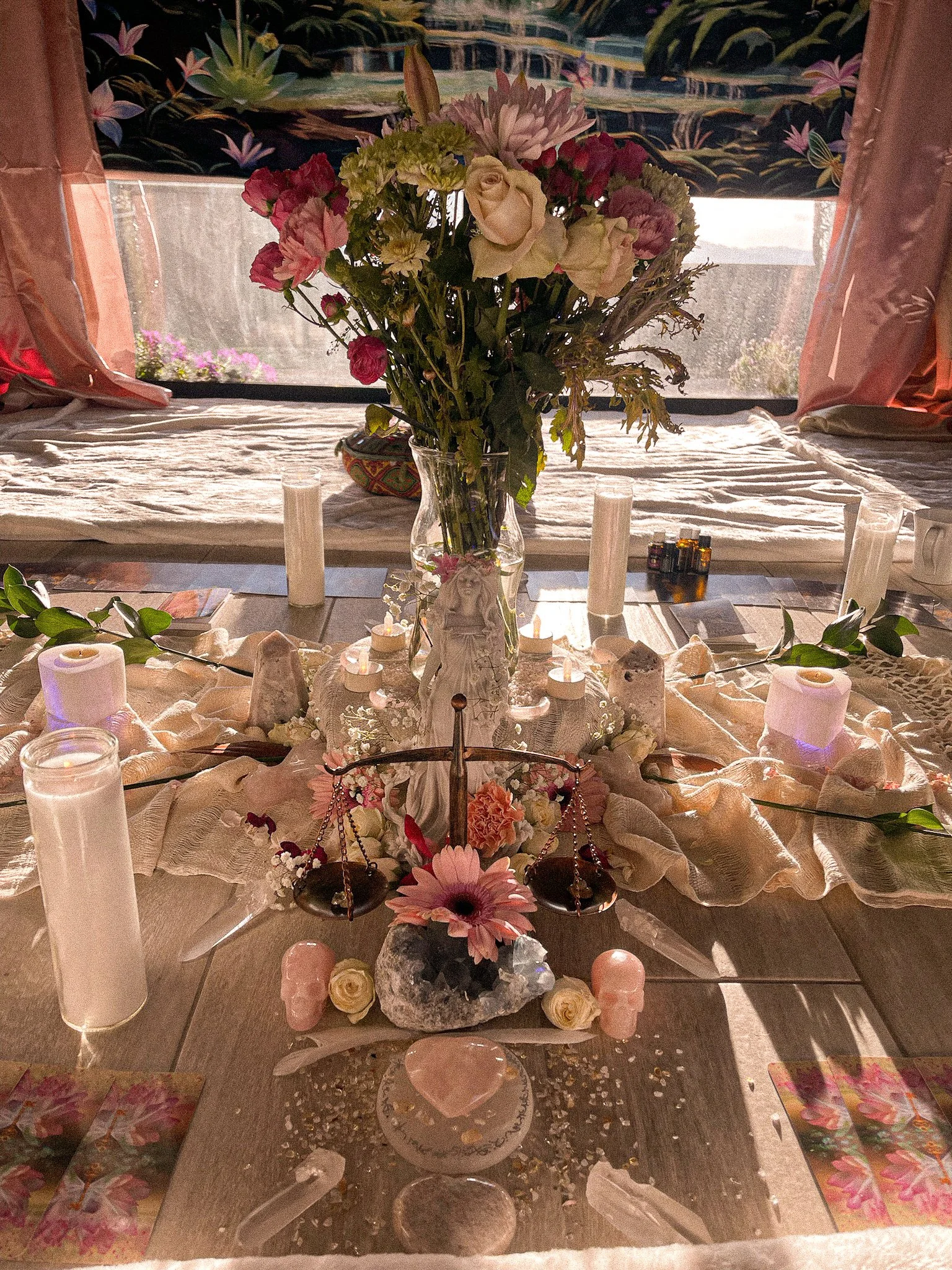 A table decorated with a large vase of mixed flowers, surrounded by candles, stones, and scales, with pink and white accents, set near a window with sunlight.