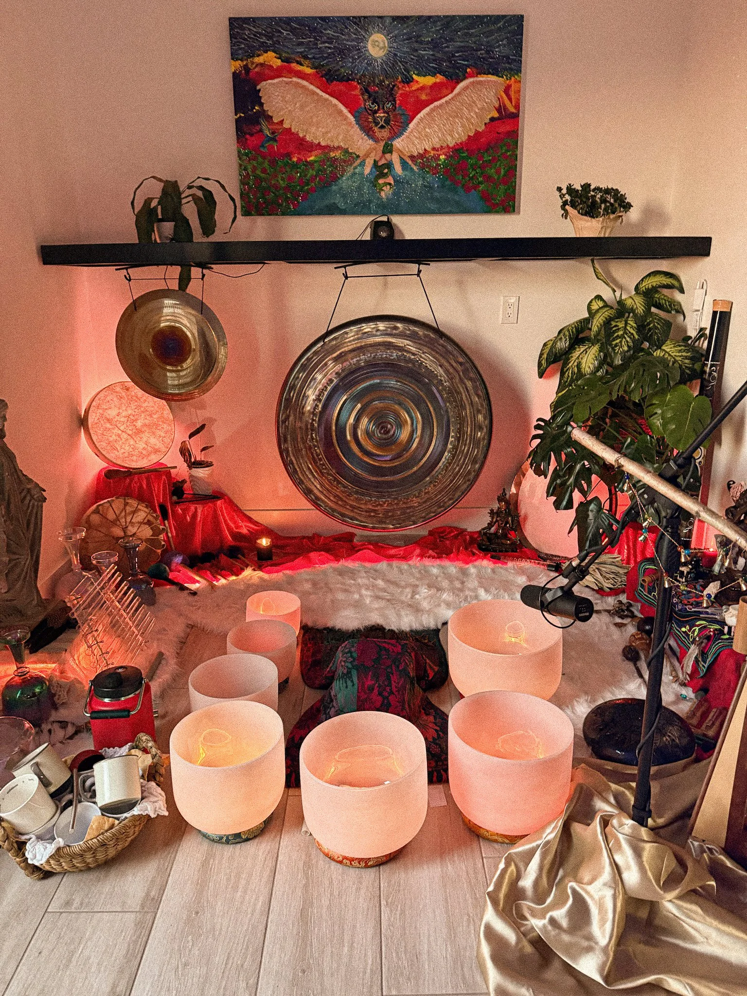 An eclectic room setup with healing and meditation items, including a large metal gong, a smaller gong, a framed artwork of a winged creature, a mounting of seven glowing singing bowls, candles, plants, and miscellaneous decor.