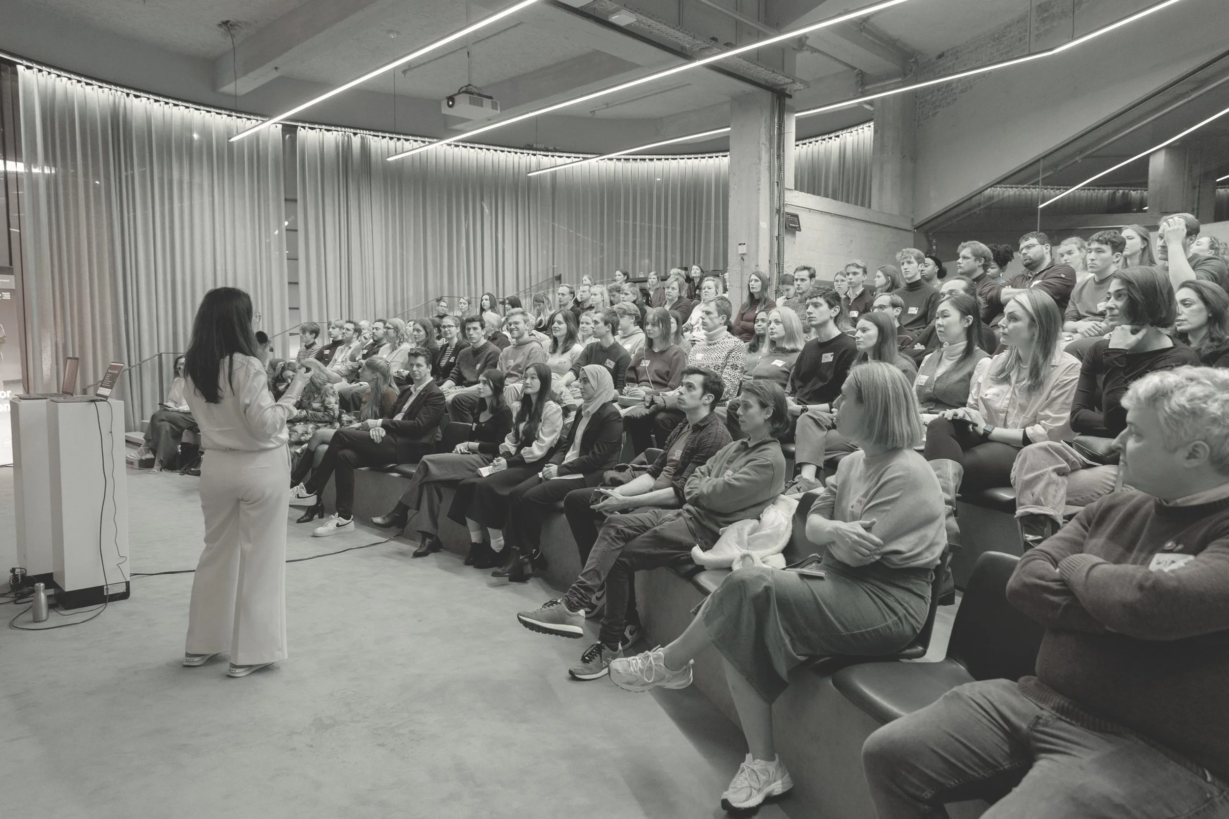A woman is giving a presentation to a large audience seated in an auditorium with modern lighting and industrial decor.