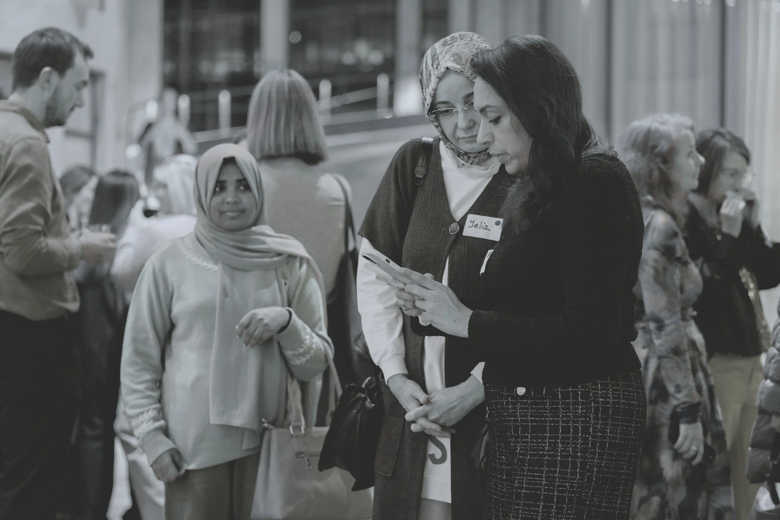 A group of diverse women and men in an indoor setting, with two women in the foreground looking at a smartphone. One woman is wearing a headscarf and a cardigan, the other has dark hair and is dressed in dark clothing. Background shows other people engaged in conversations.