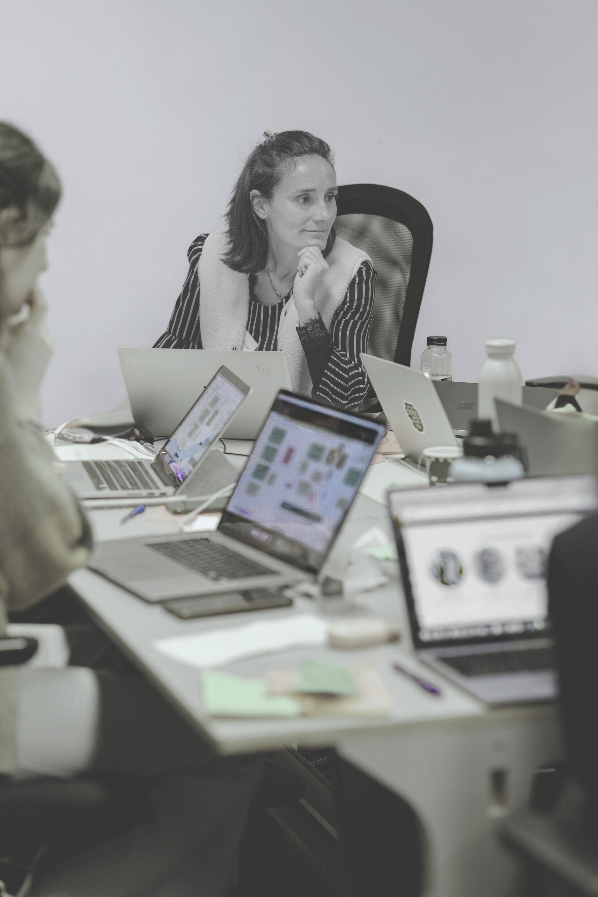 A woman sitting at a conference table with several laptops, water bottles, and notes, appearing focused during a meeting.