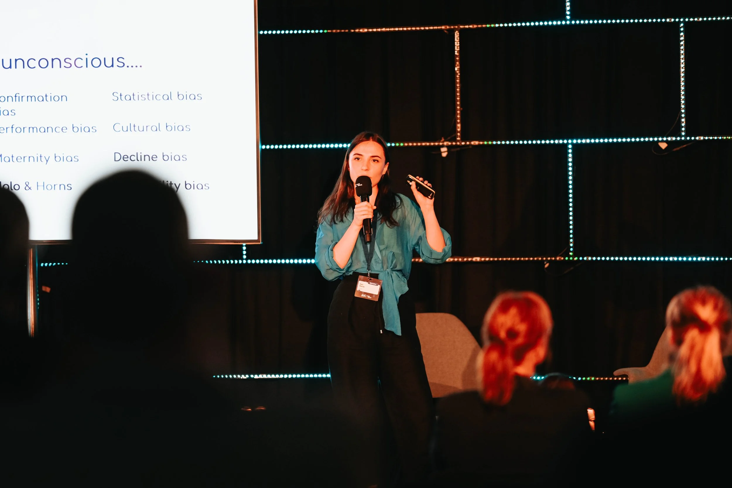 A young woman presenting at a conference on stage, holding a microphone and a remote control, with a large screen behind her displaying a slide on unconscious biases.