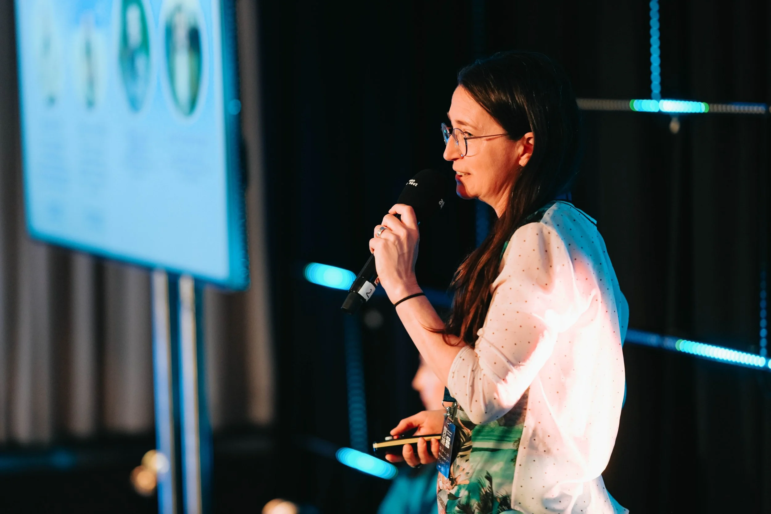 A woman with glasses and dark hair is speaking into a microphone on stage with a large screen showing a presentation behind her.