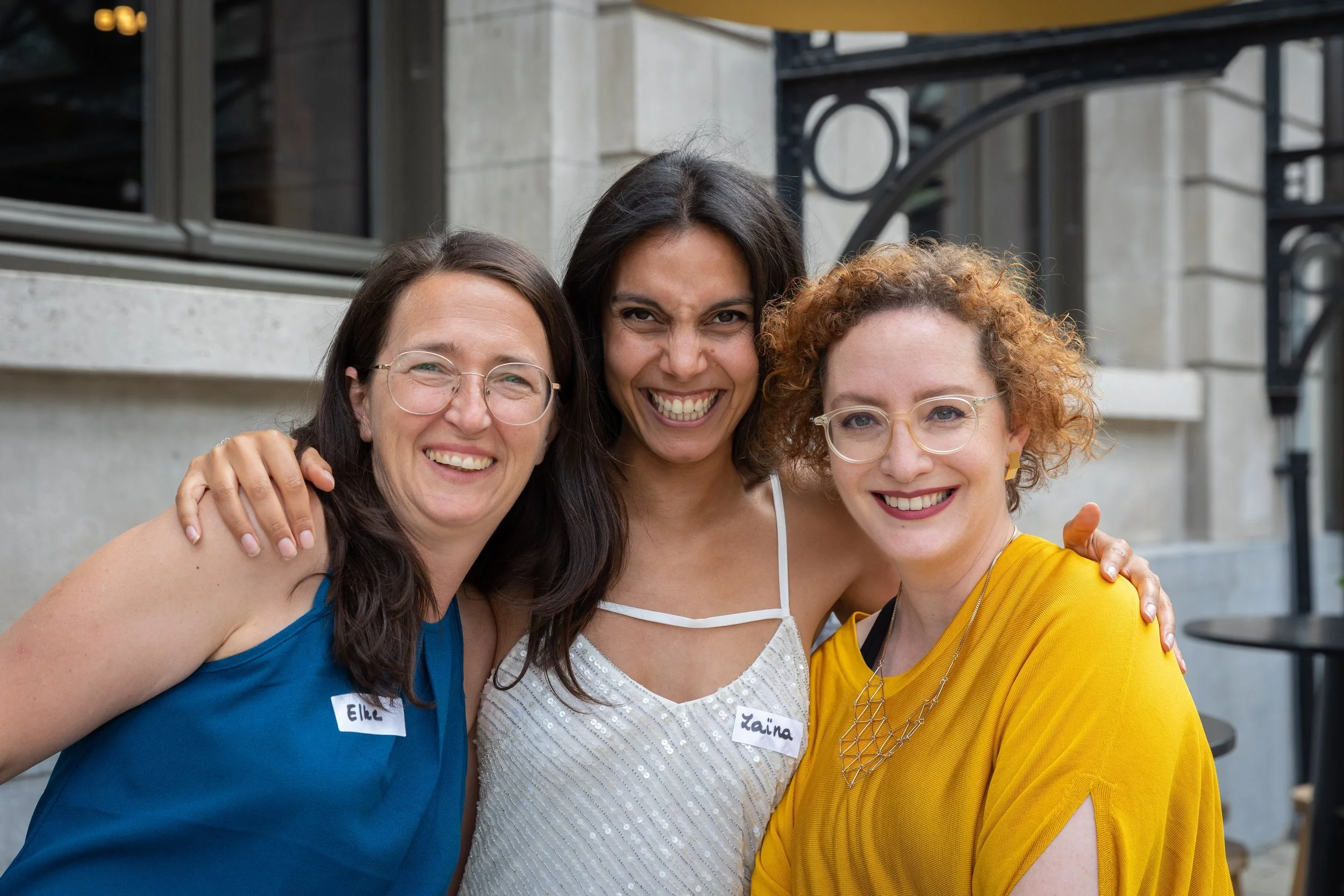 Three women smiling and posing together outdoors, wearing colorful tops, with name tags reading Elle, Zaina, and an unreadable third name.