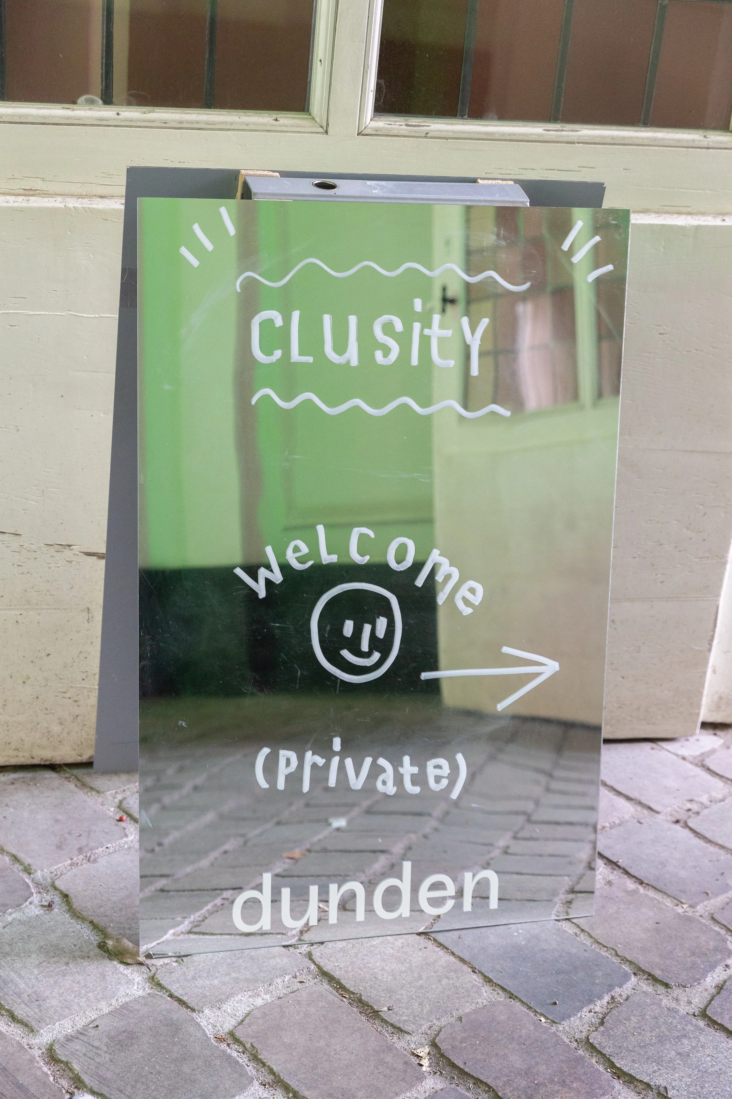 A glass sign on a sidewalk with the words 'Clusity', 'welcome', '(private)', and 'dunden', with a smiley face and a right arrow. The background shows a building and a window.