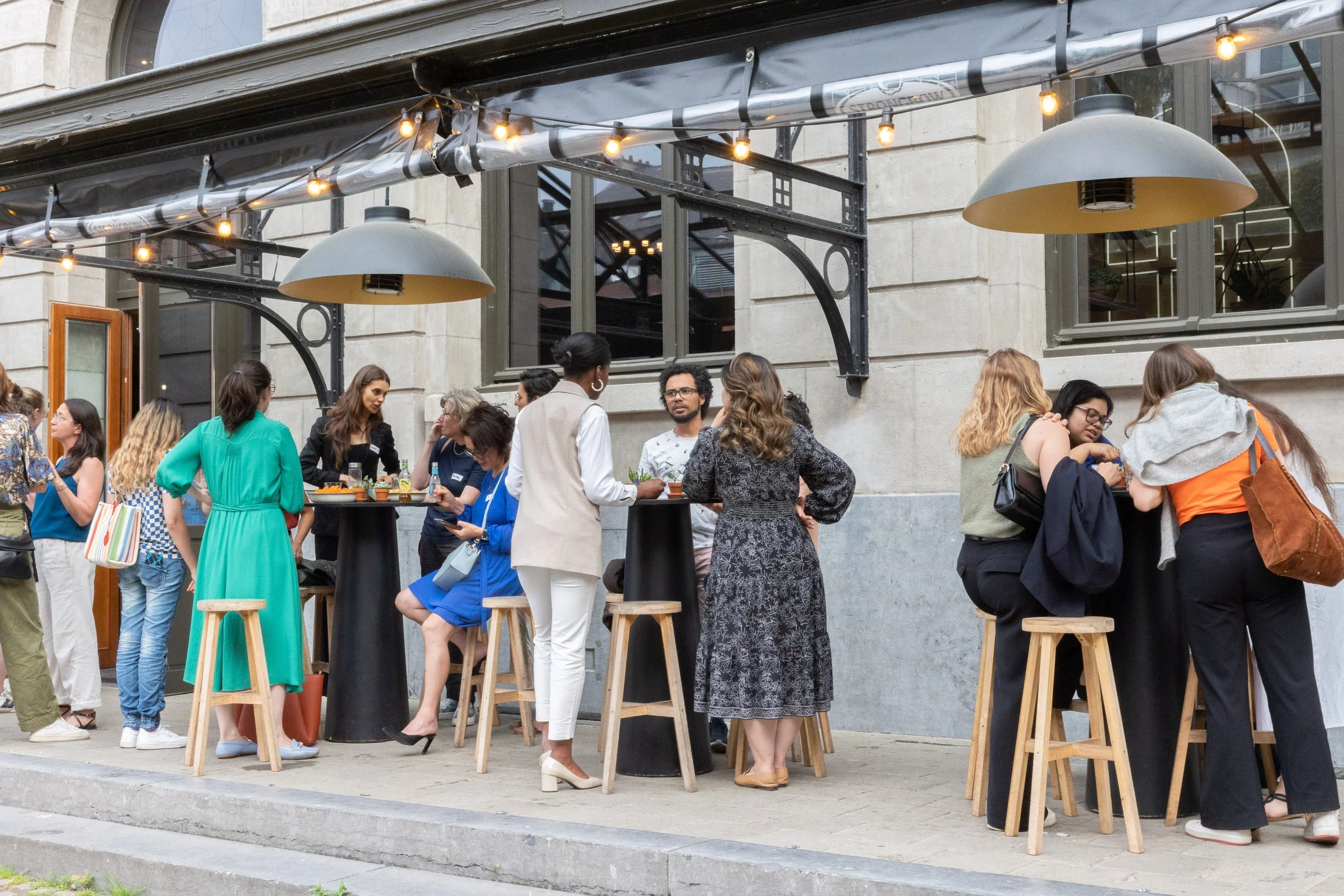Group of diverse people socializing at an outdoor bar or cafe on a city sidewalk during daytime.