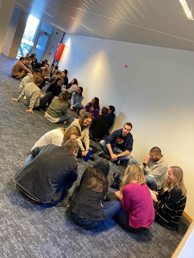 Group of people sitting on the floor in a corridor, engaged in conversation.