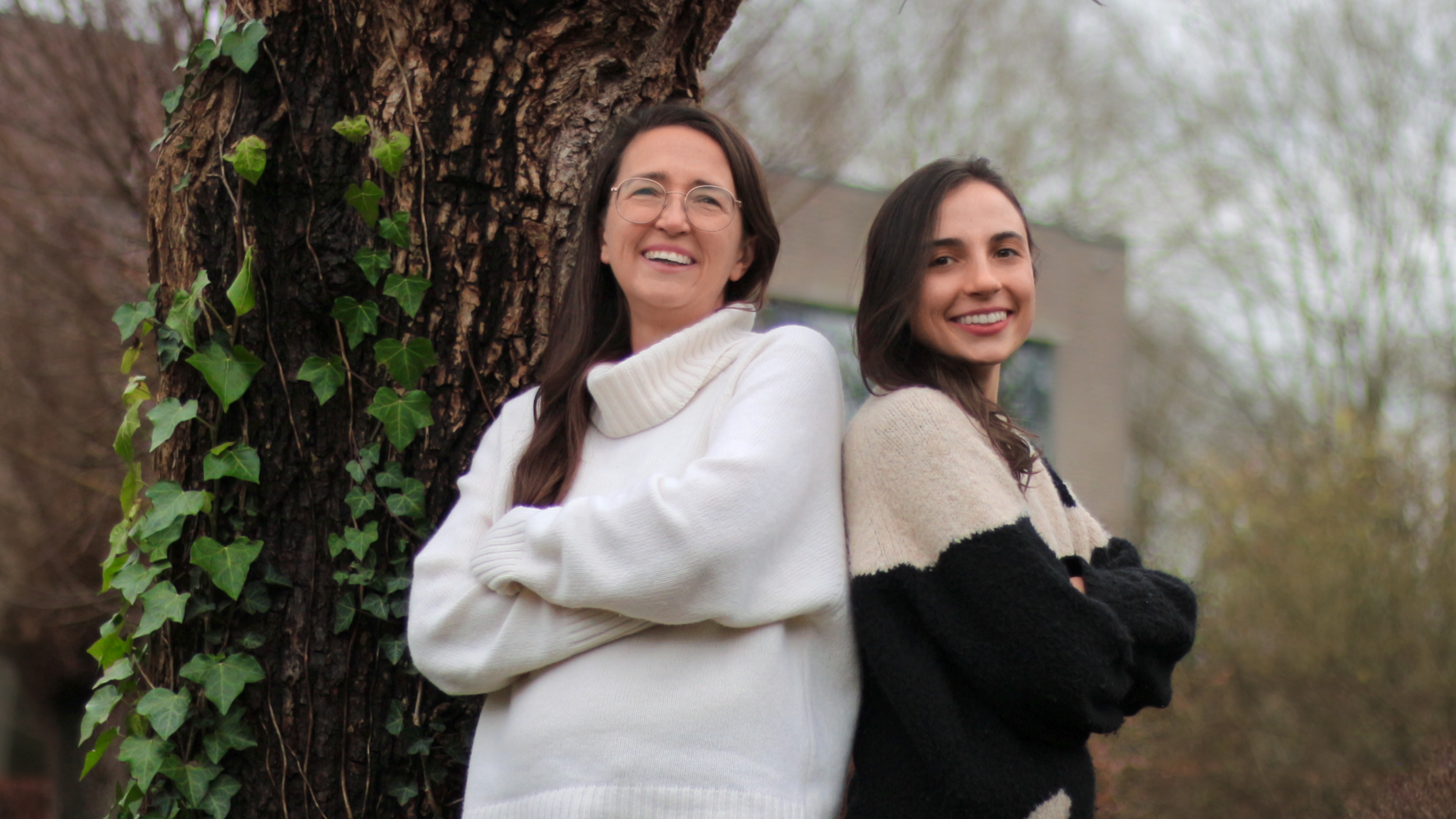 Two women standing outdoors near a tree with ivy, smiling and leaning against each other.