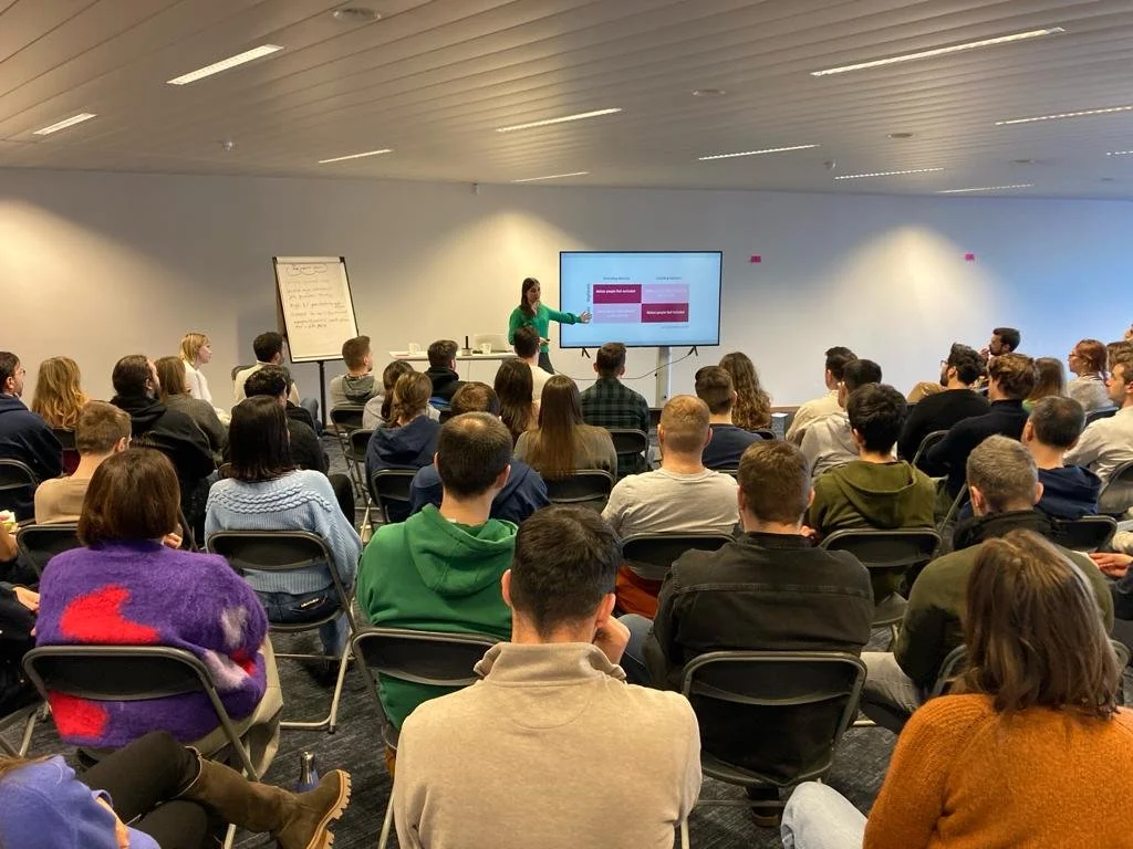 A woman giving a presentation to a large group of people seated in an indoor conference room with a screen and a flip chart.