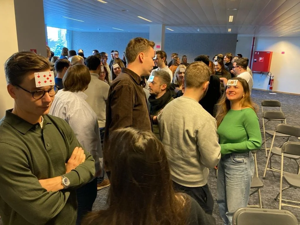 Group of young adults at an indoor event, with some participating in a game or activity, wearing stickers on their foreheads, socializing, and standing in a casual setting.