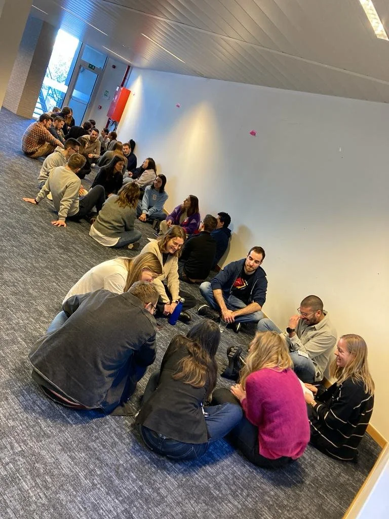 Group of people sitting on the floor in a corridor, engaged in conversation.