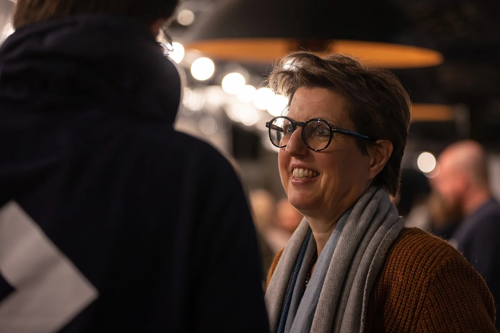 A woman with short brown hair, glasses, and a gray scarf smiling and talking to a person in a black hoodie in a crowded indoor setting.