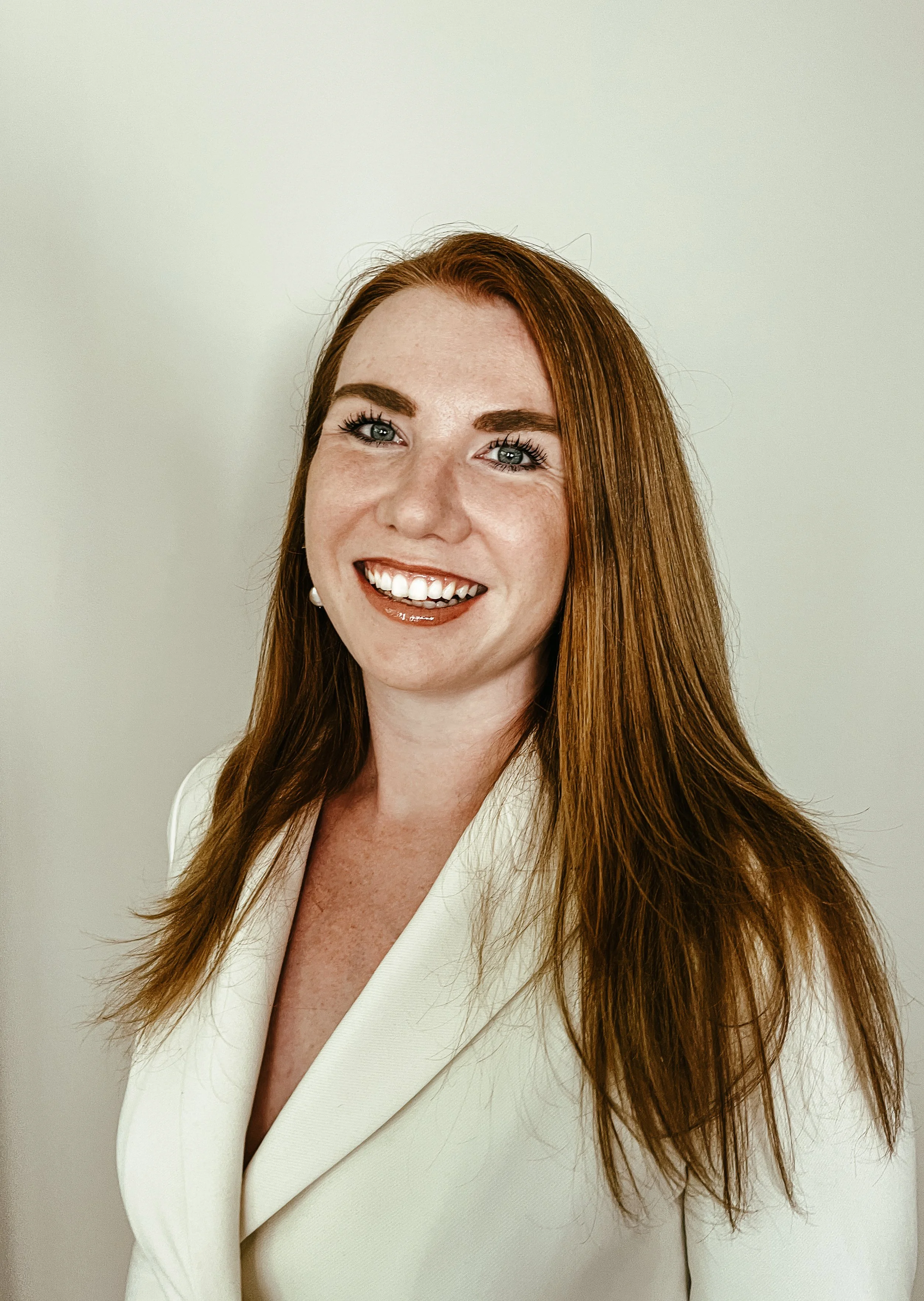 A woman with long red hair, blue eyes, and a white blazer smiling against a plain light background.