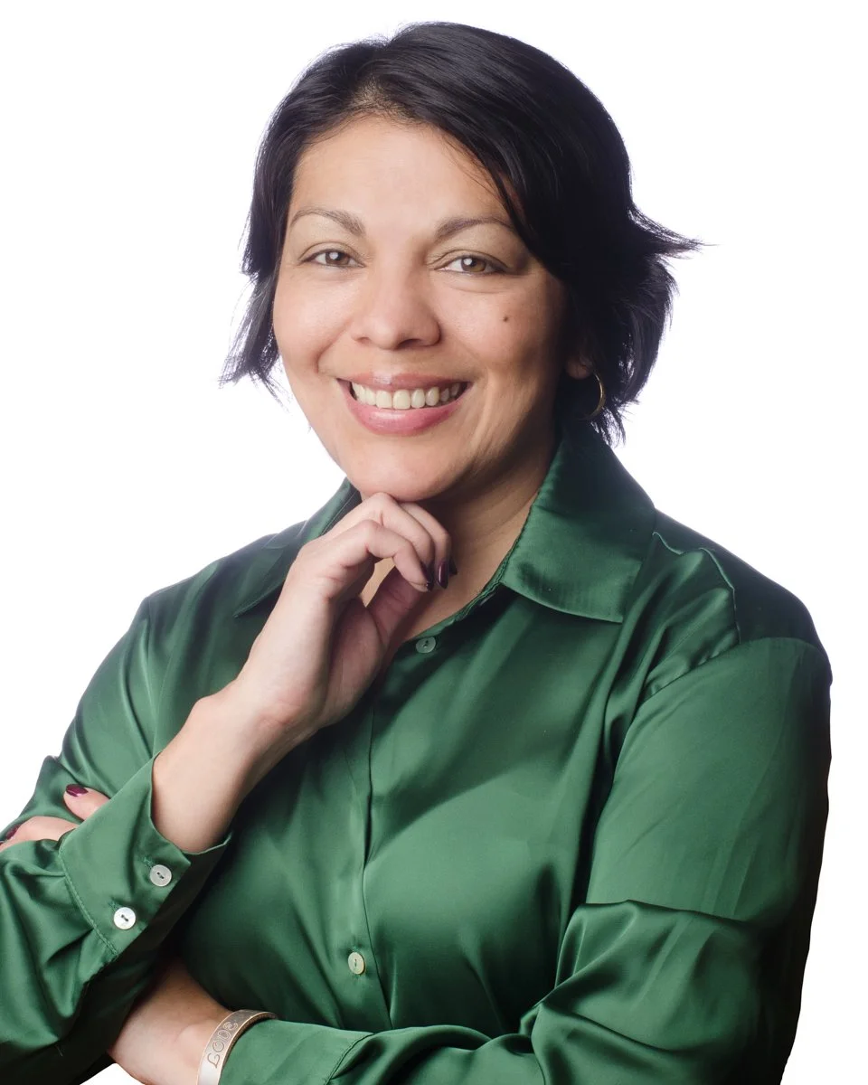 A woman with dark hair styled in a short bob, smiling, wearing a shiny green satin shirt, posing with one hand resting on her chin and the other crossed over her chest against a white background.