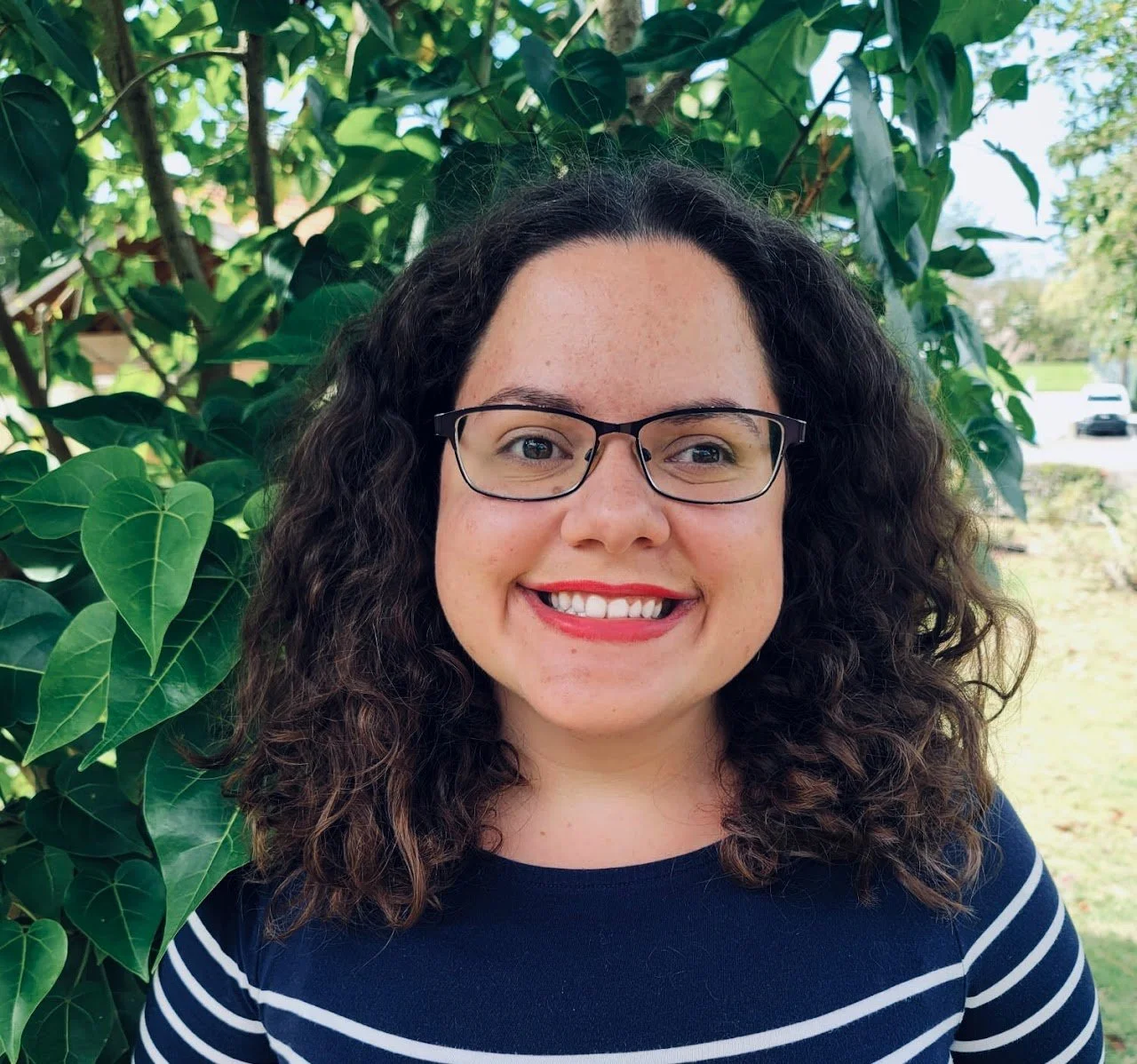 A woman with curly hair wearing glasses smiling outdoors near green leafy plants.