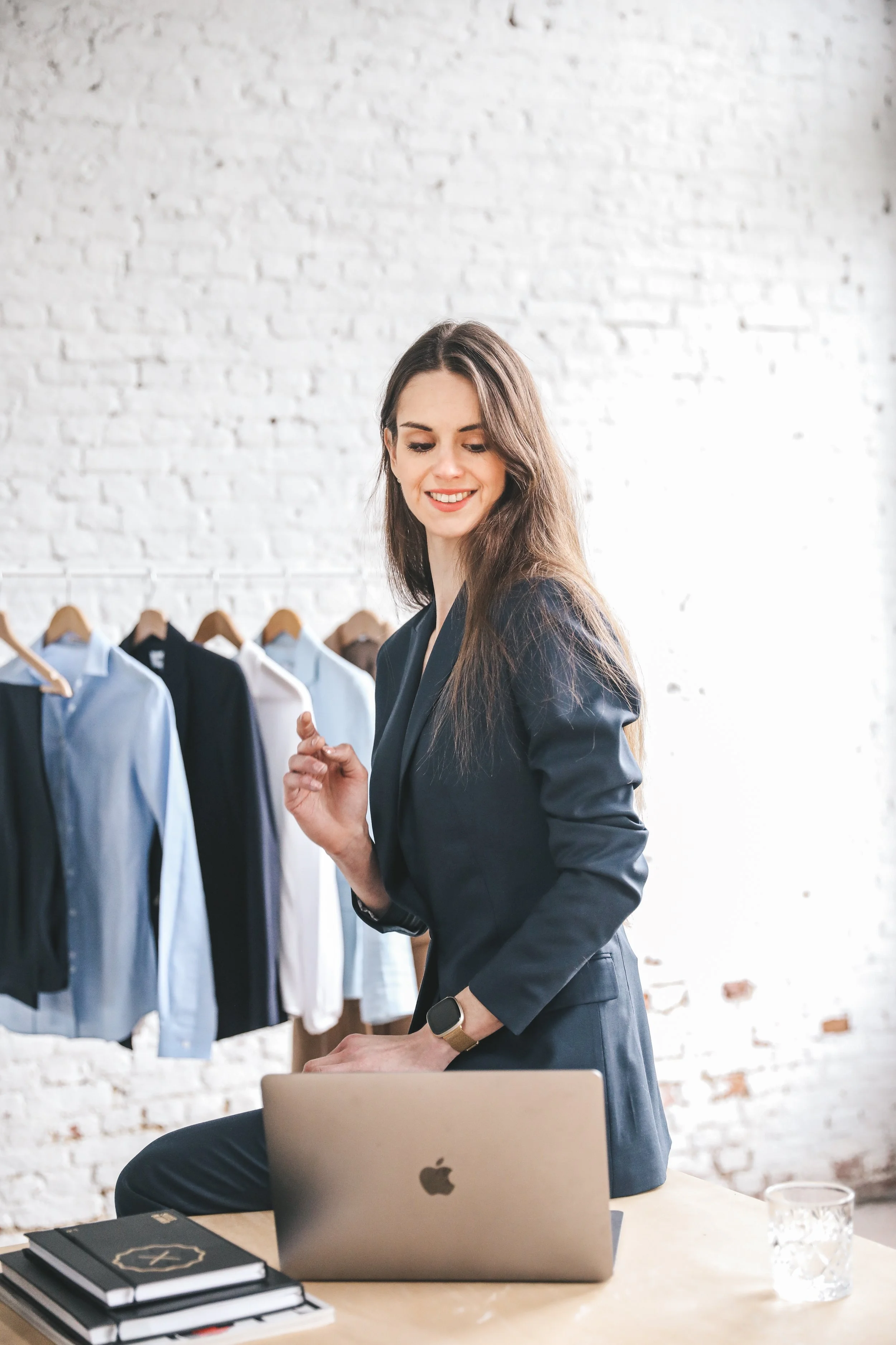 A woman with long brown hair working on a silver MacBook in a modern office space with a white brick wall and clothing rack behind her.